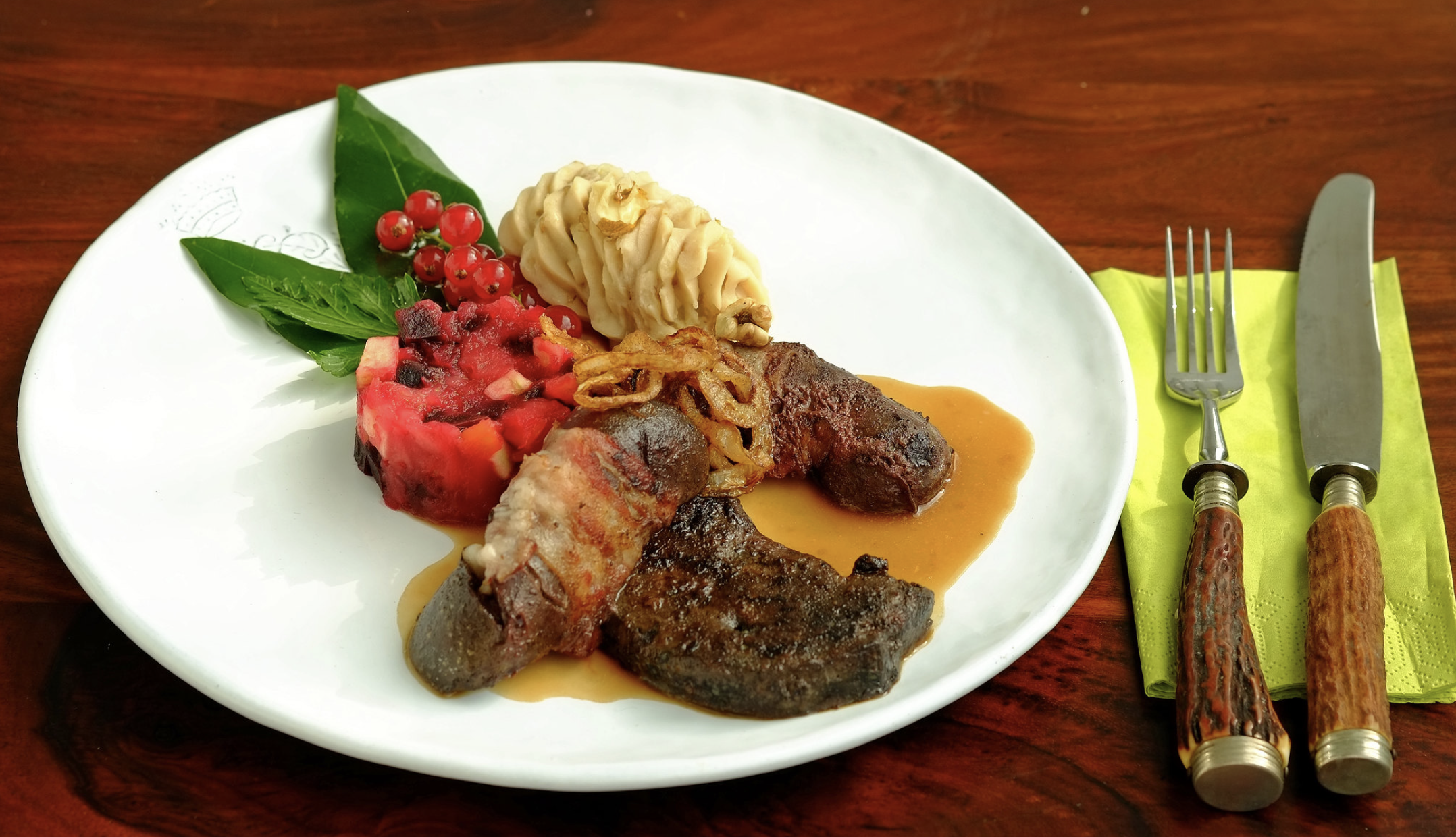 A white plate with cooked meat in brown sauce, mashed potatoes, a slice of berry terrine, red currants, green leaves, and crispy onions. Knife and fork rest on a yellow napkin on a wooden table.