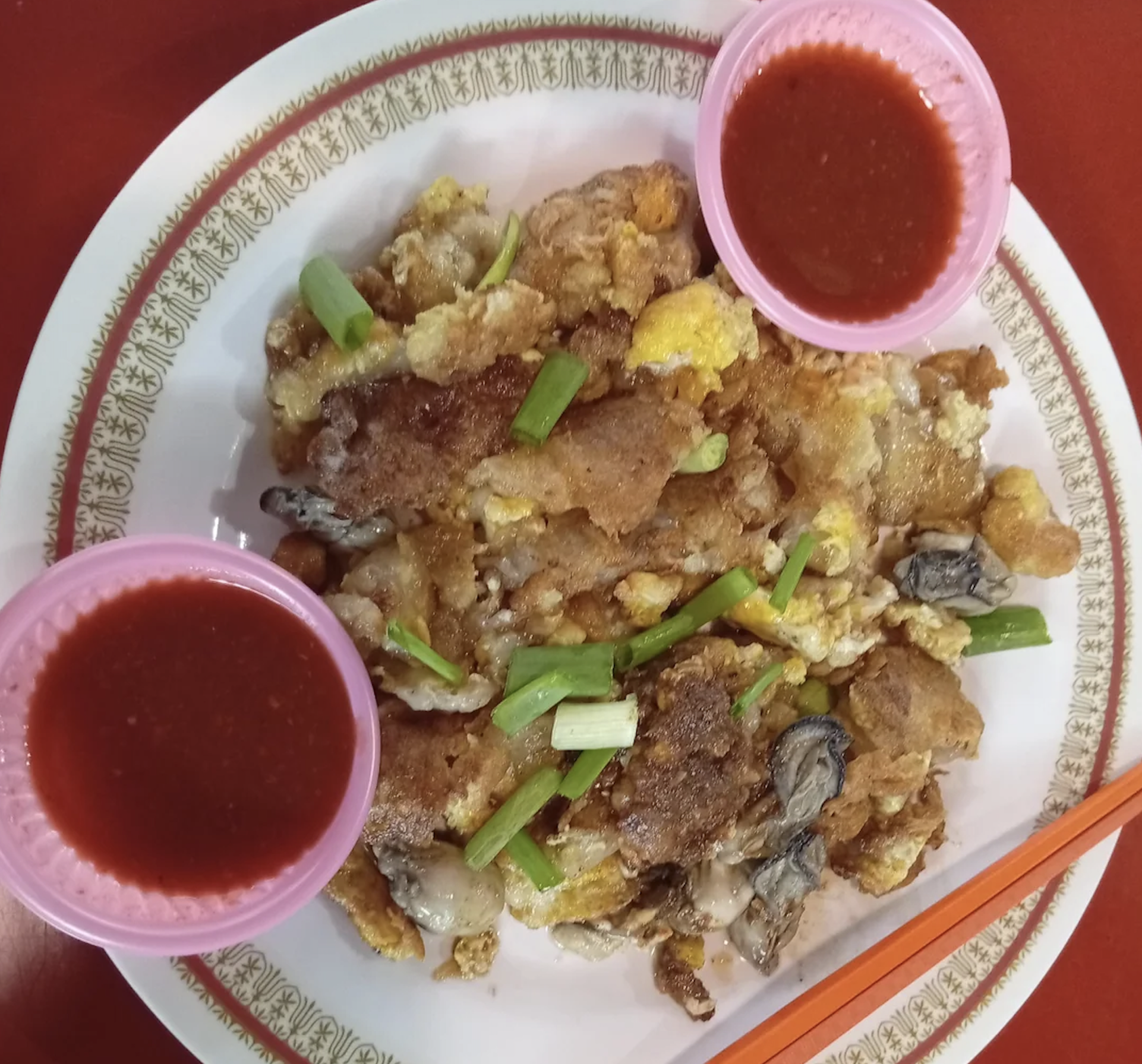 A plate of fried oyster omelette garnished with green onions, served with two small bowls of red sauce, on a patterned plate with orange chopsticks partially visible.