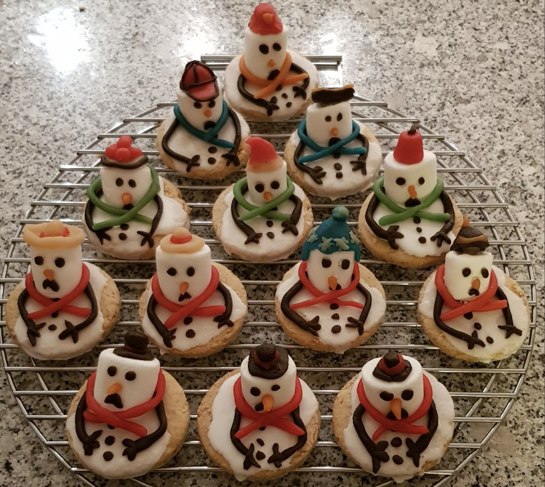 Snowman-shaped cookies decorated with white icing, colored scarves, hats, and chocolate details are arranged in a triangle shape on a wire rack over a granite countertop.