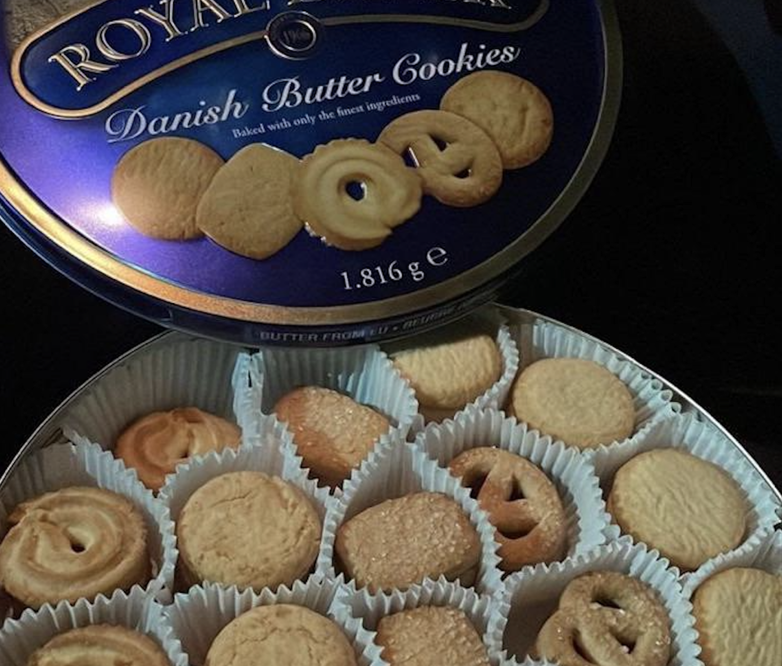 An open blue tin of Danish butter cookies shows assorted cookie shapes arranged in white paper cups, with the lid resting behind the tin.
