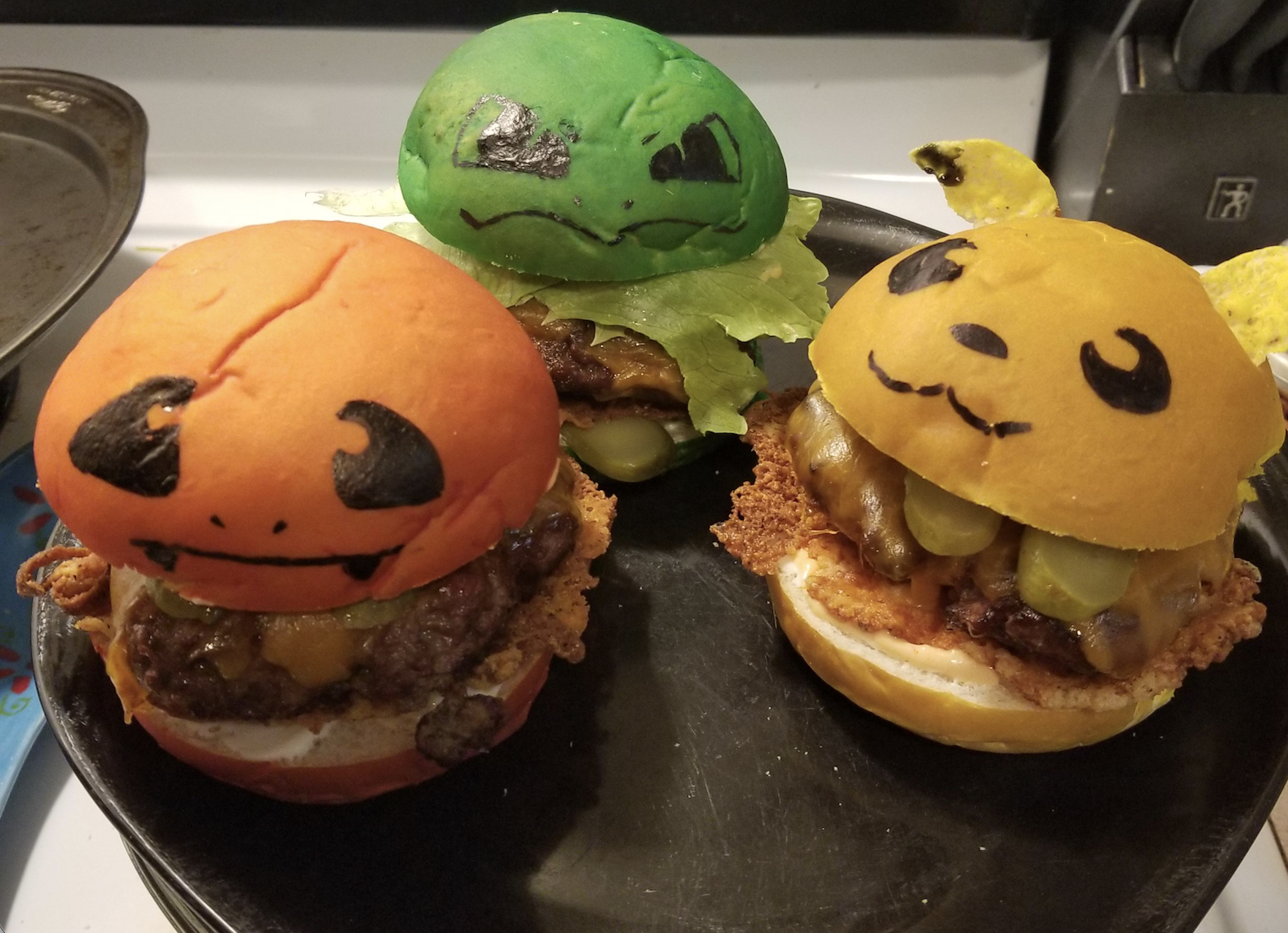 Three cheeseburgers with buns decorated and colored to look like Pokémon characters: Charmander (orange), Bulbasaur (green), and Pikachu (yellow), on a black plate.
