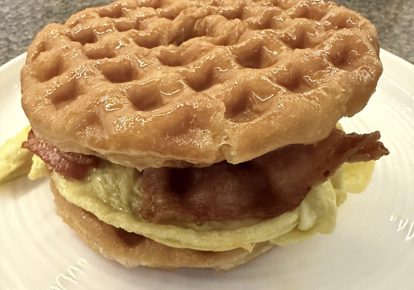 A breakfast sandwich with bacon and egg between two waffle halves, drizzled with syrup, sits on a white plate.