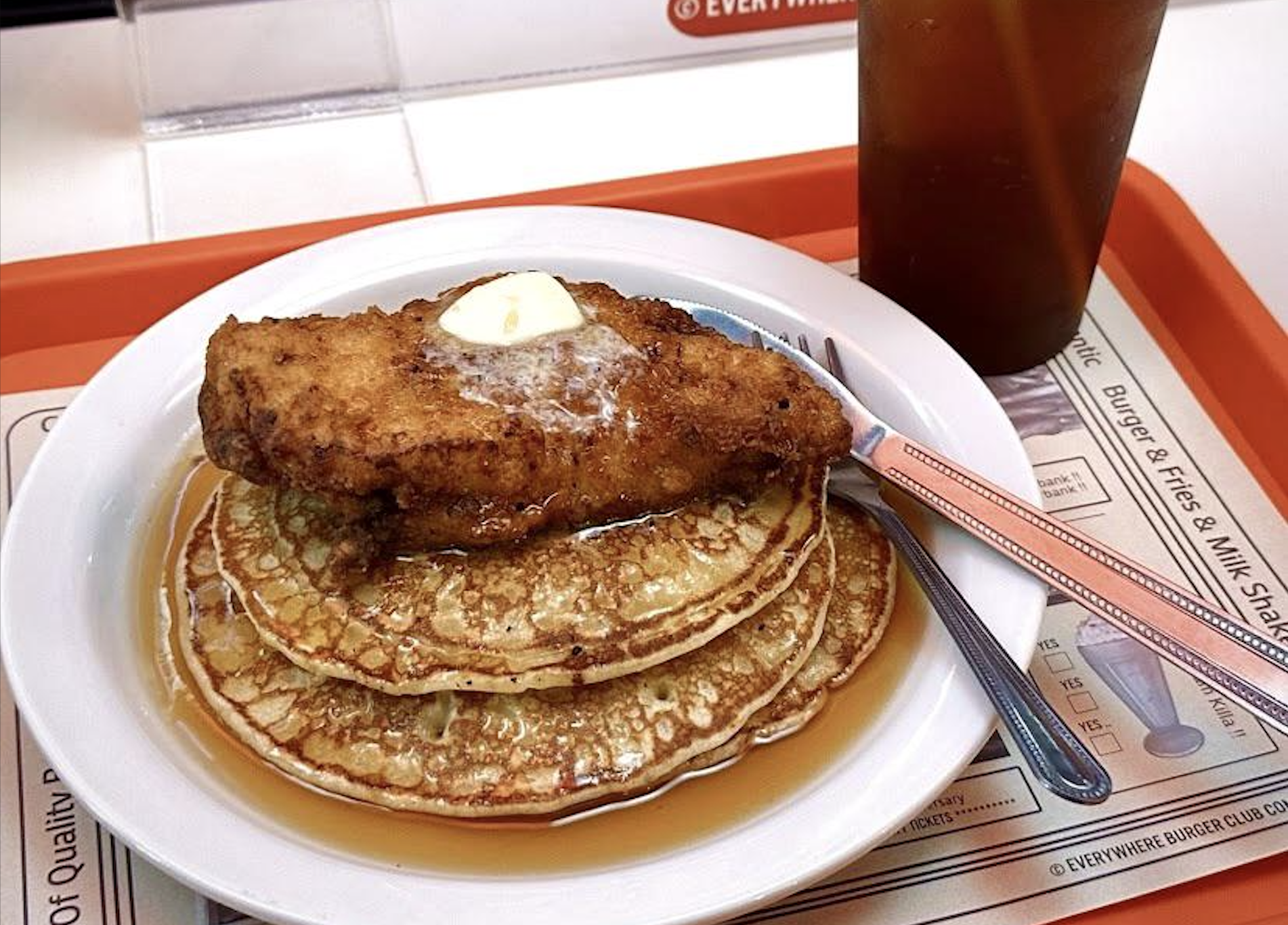 A plate of pancakes topped with a piece of fried chicken and a pat of butter, covered in syrup, with a fork and knife on the side and a glass of iced tea in the background.