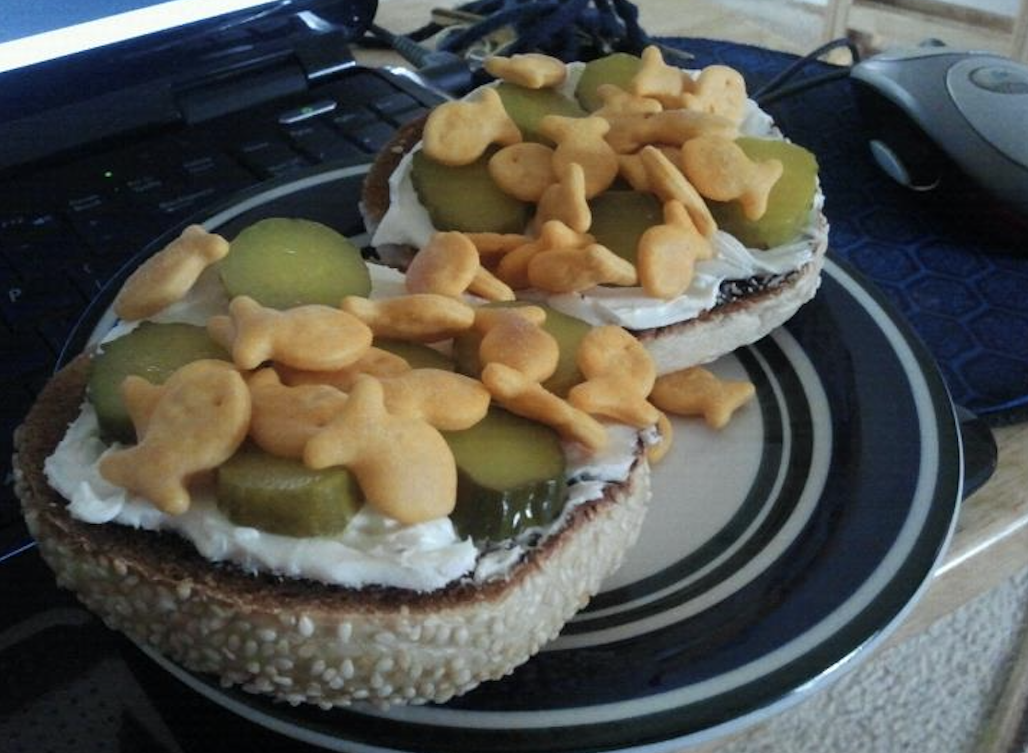 An open sesame bagel with cream cheese, pickle slices, and cheddar Goldfish crackers on a plate, next to a laptop and computer mouse.