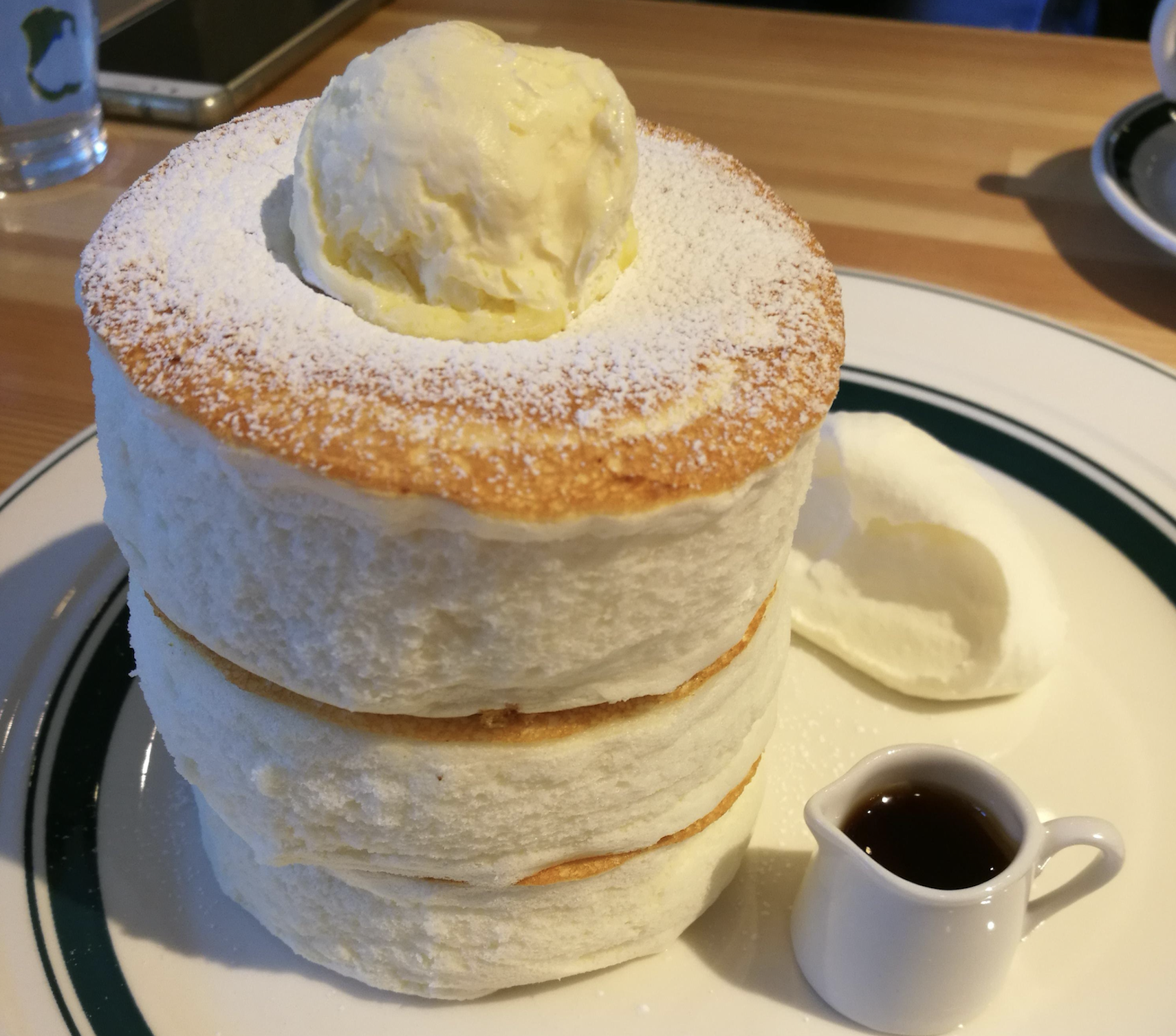 A stack of fluffy Japanese souffle pancakes topped with a scoop of butter or cream, dusted with powdered sugar, served with a small pitcher of syrup and a dollop of whipped cream on the side.