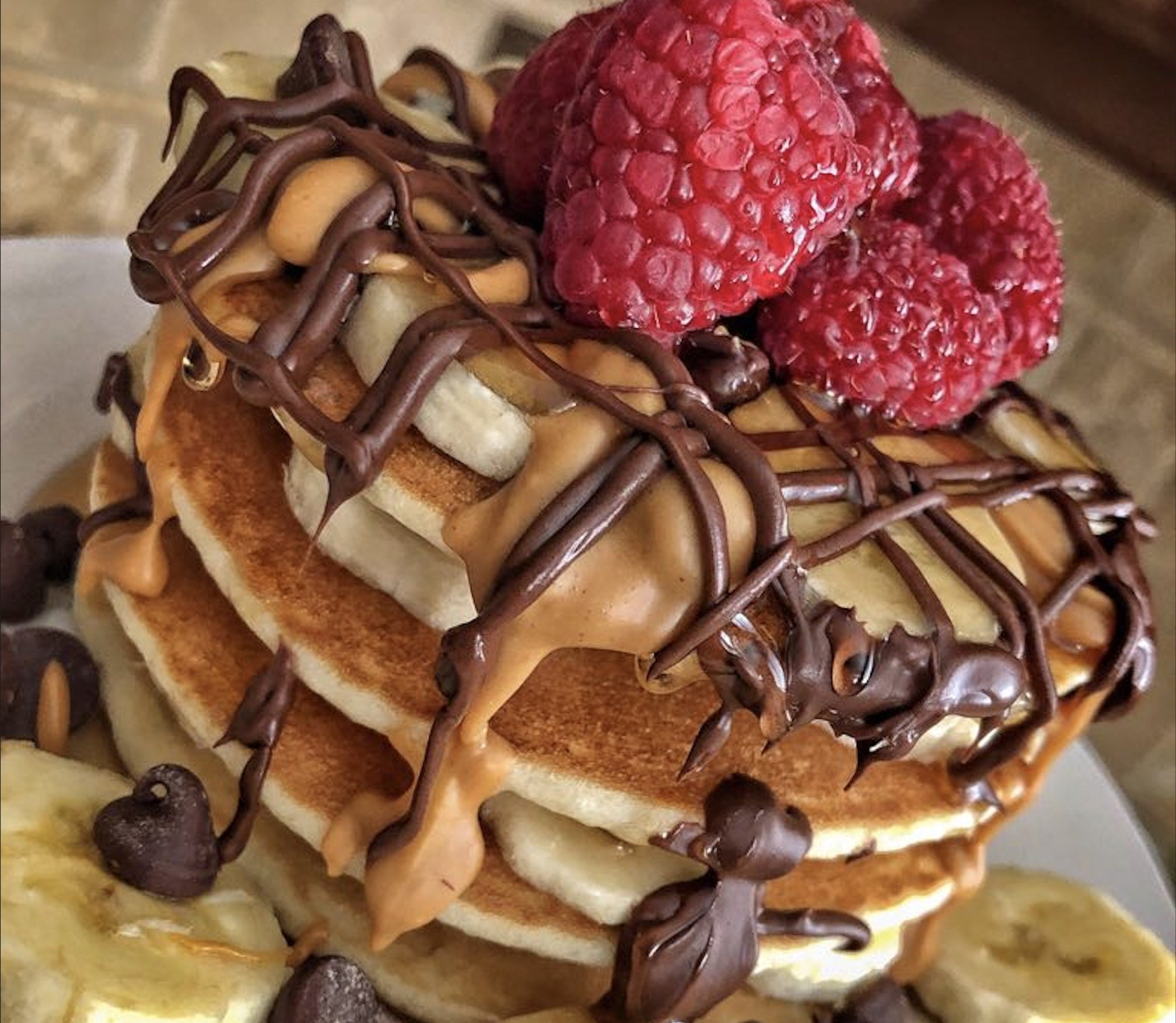 A stack of pancakes topped with sliced bananas, raspberries, chocolate chips, and drizzled with chocolate and peanut butter sauce.
