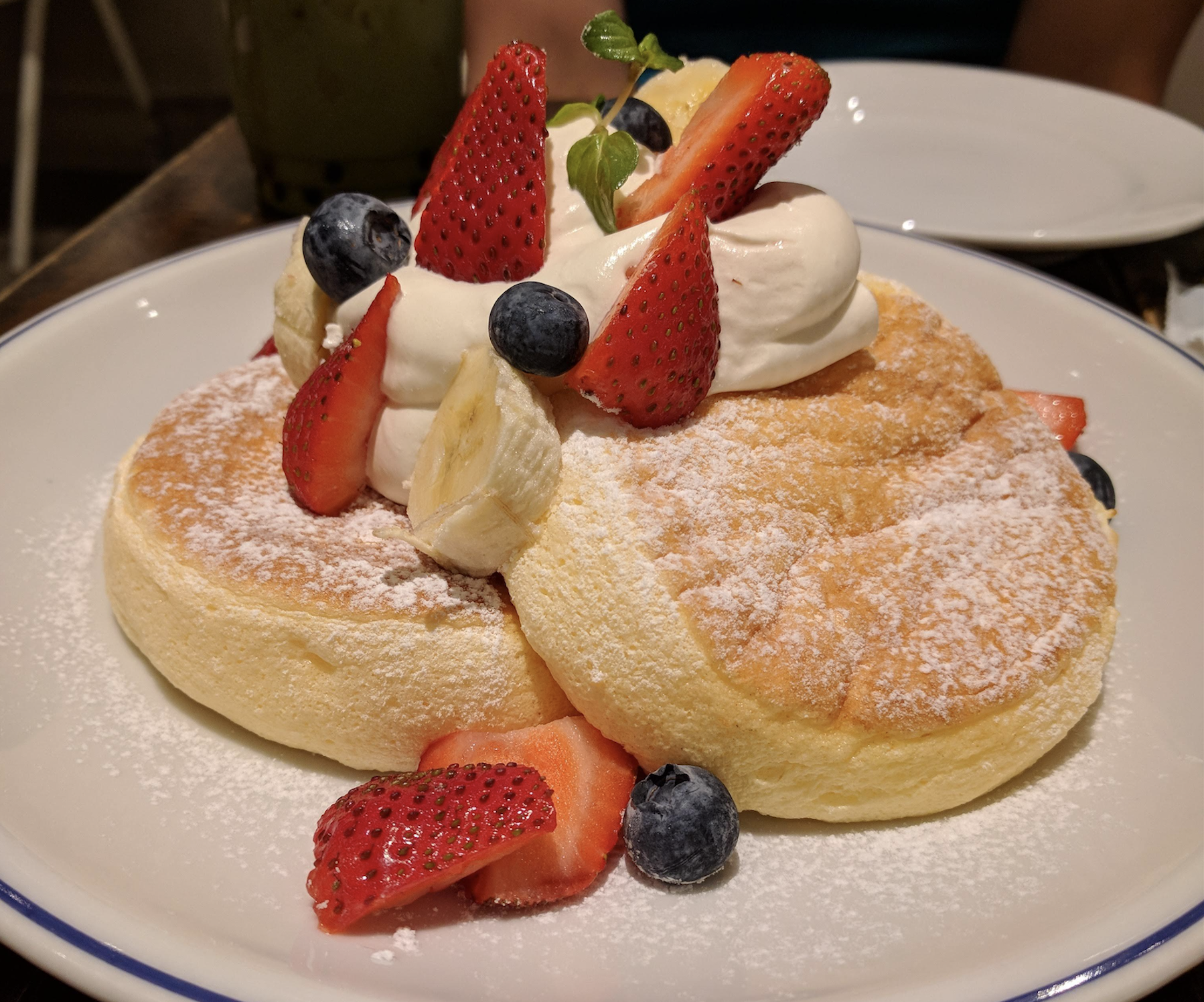 A plate of fluffy pancakes topped with whipped cream, sliced strawberries, blueberries, banana, and powdered sugar, served on a white plate.