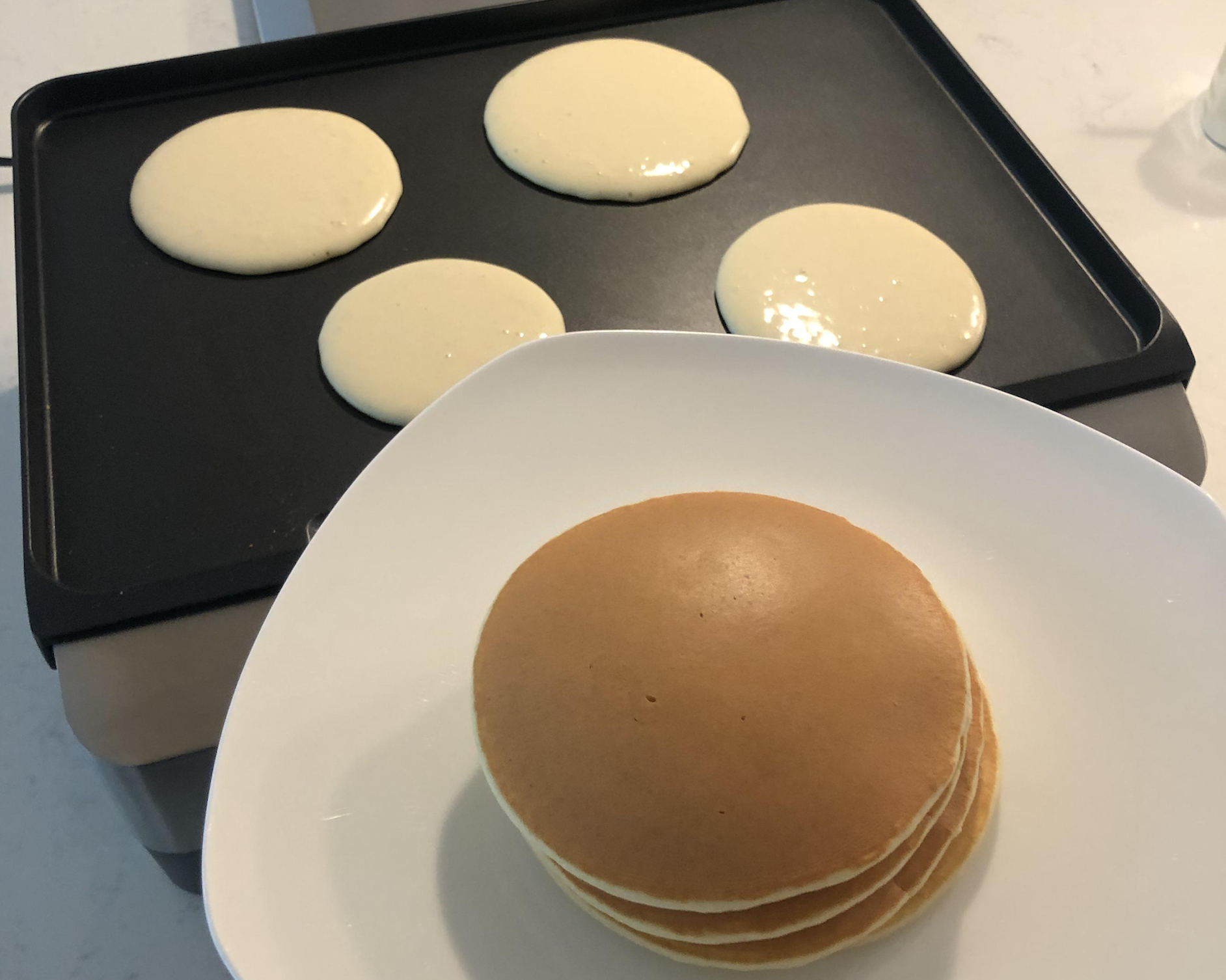 A stack of golden pancakes sits on a white plate in the foreground, while four pancakes cook on a black griddle in the background.