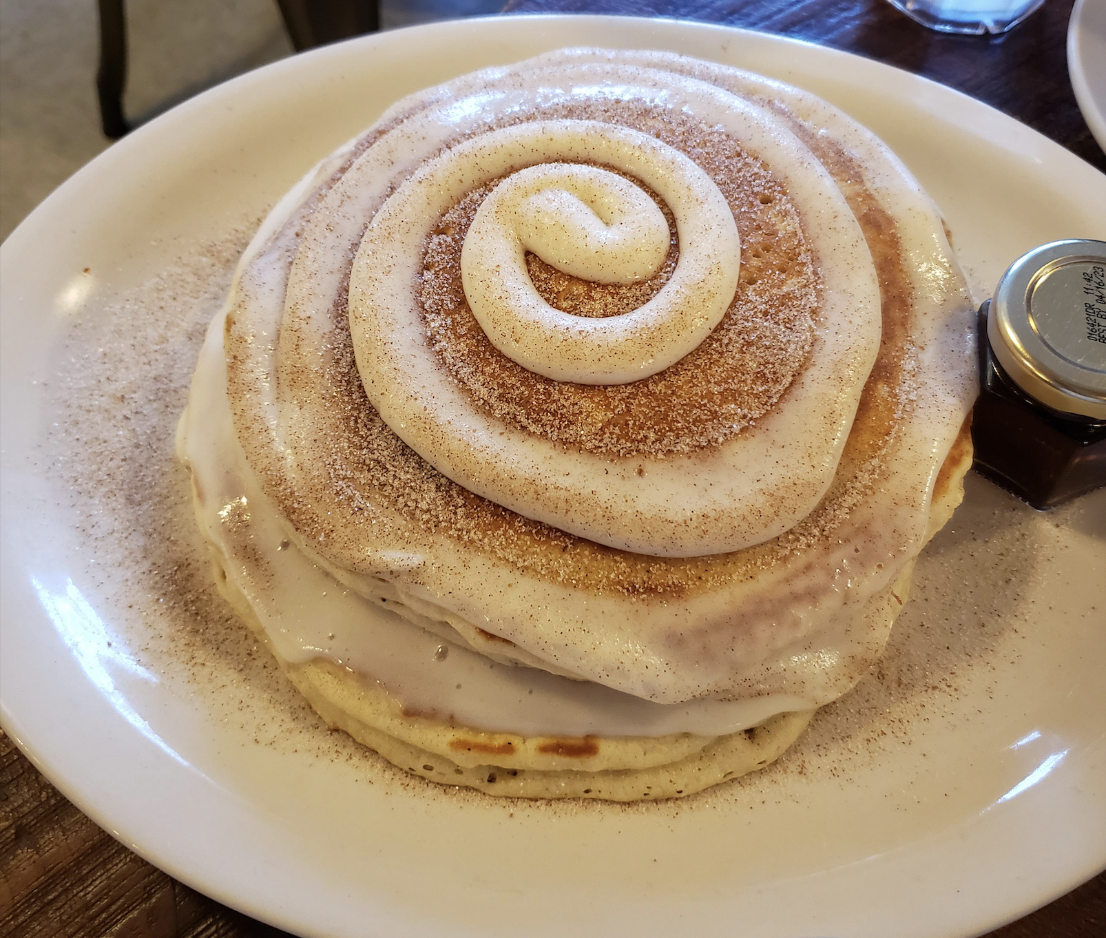 A stack of pancakes topped with icing and a swirl of cinnamon, served on a white plate with a small jar of syrup on the side.