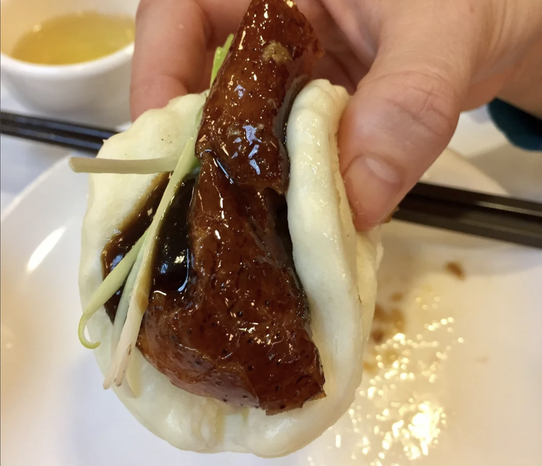 A hand holds a Chinese bao bun filled with glazed roasted duck, sauce, and green onions. A pair of chopsticks and a bowl with yellow liquid are in the background.