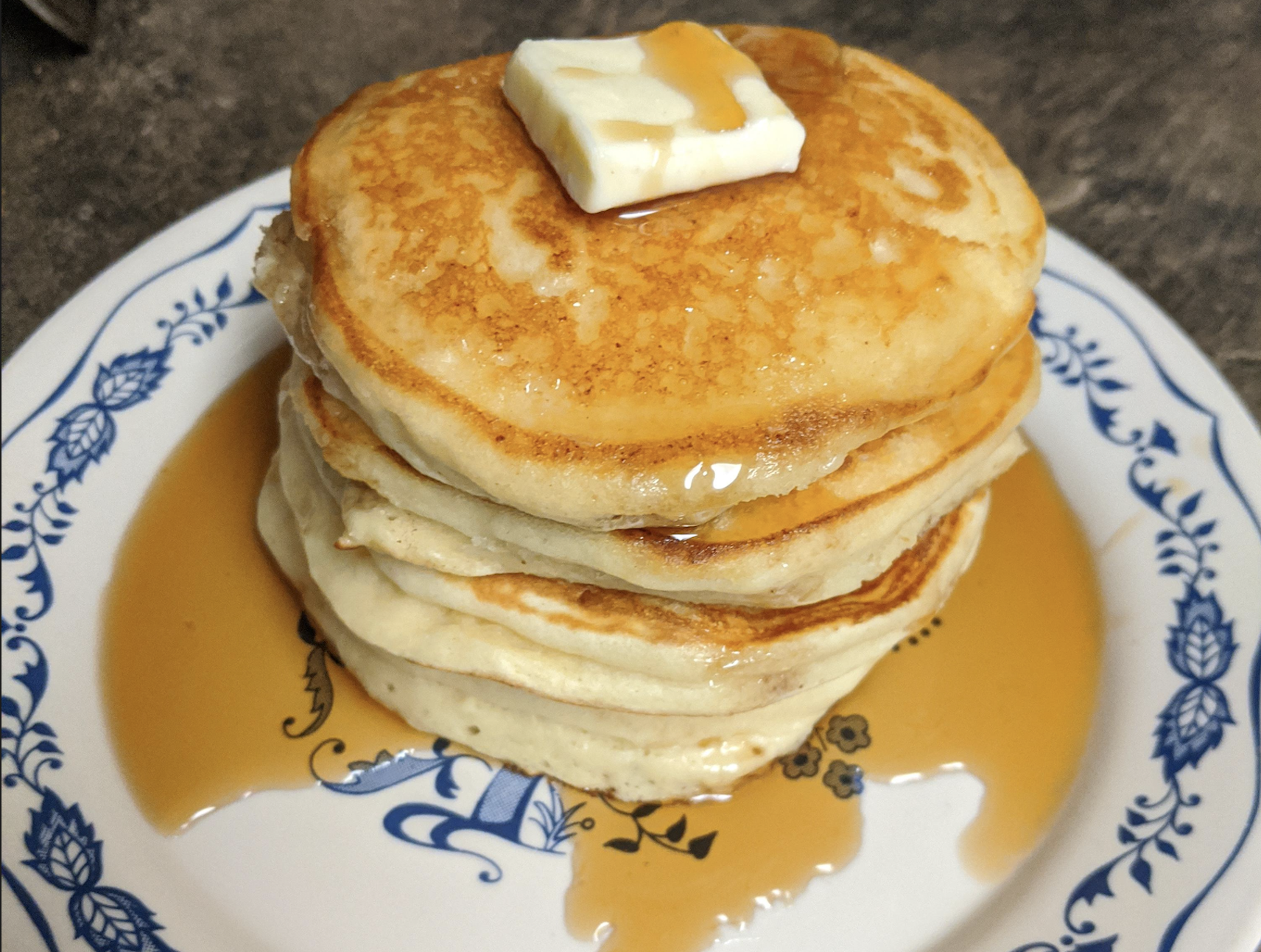 A stack of fluffy pancakes topped with a pat of butter and drizzled with syrup sits on a white plate with blue floral designs.