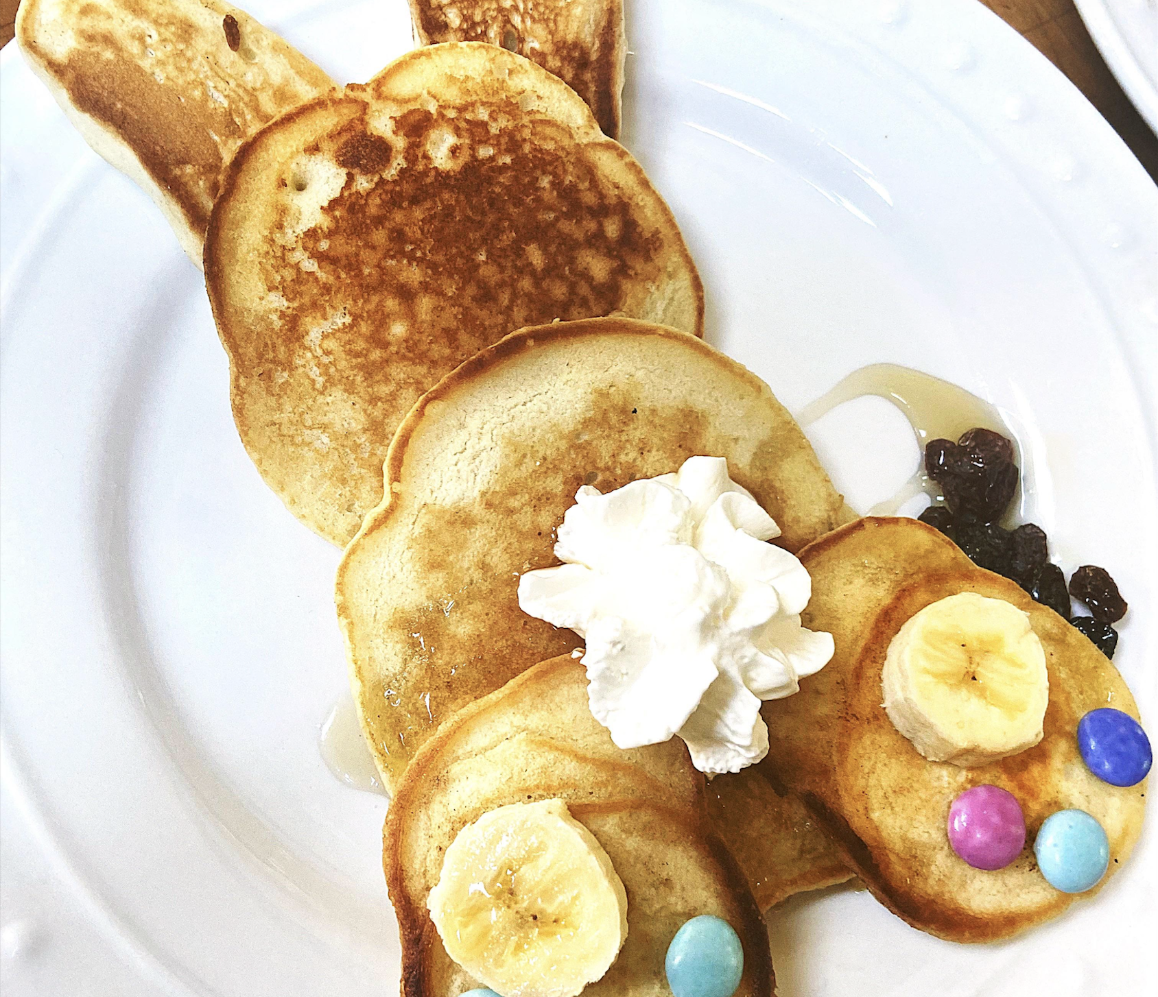 Three pancakes arranged like a bunny, with banana slices, colorful candies, whipped cream as a tail, and syrup on a white plate.