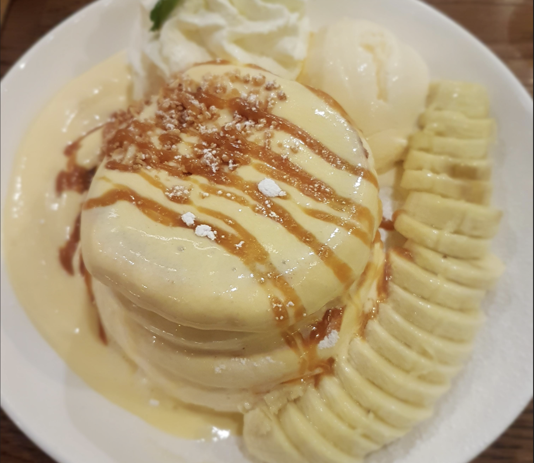A plate with a fluffy pancake drizzled with caramel sauce, served with whipped cream, a scoop of vanilla ice cream, and sliced bananas arranged on the side.