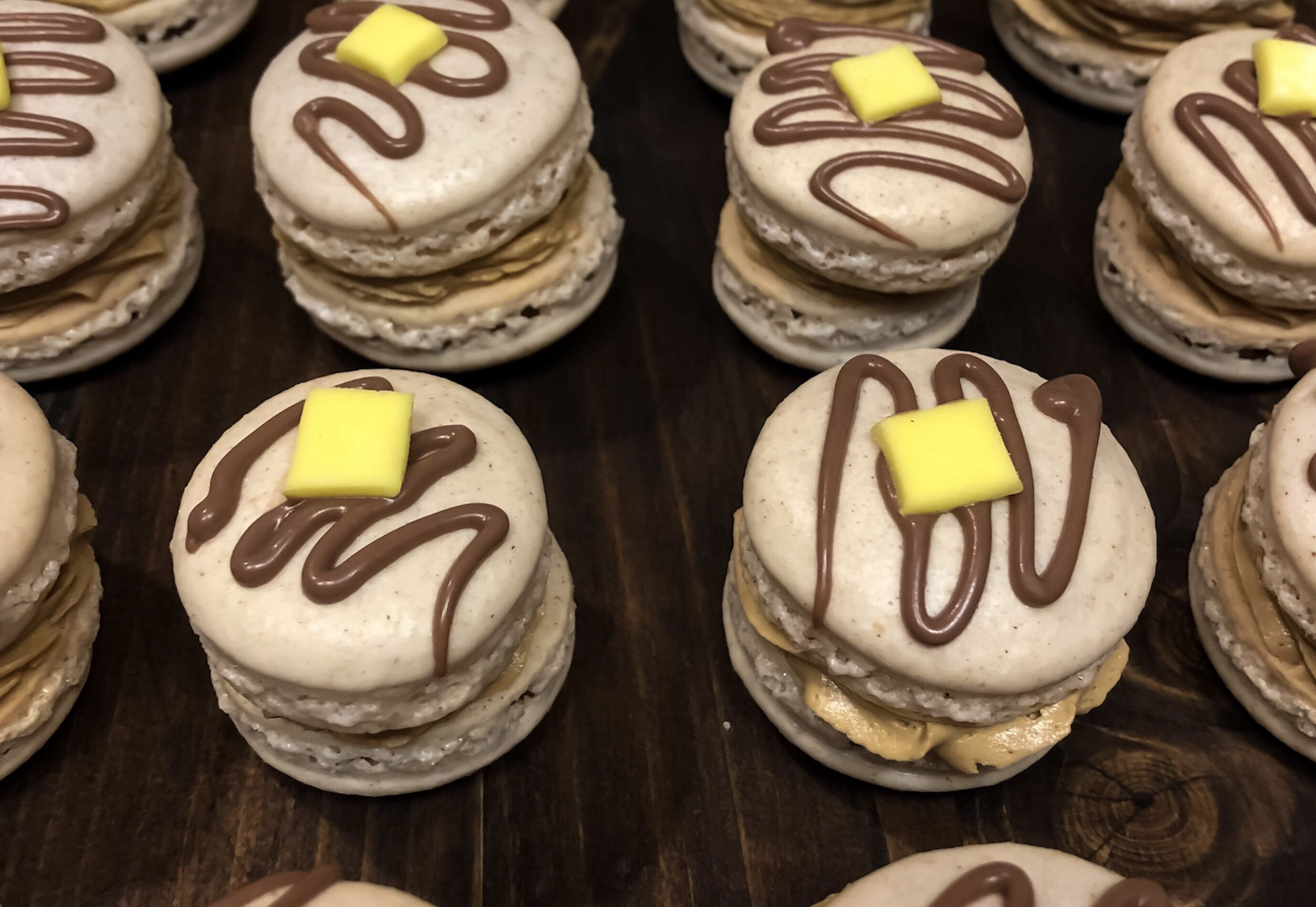 Macarons with tan filling, drizzled with chocolate, and topped with a small yellow square, arranged on a dark wooden surface.