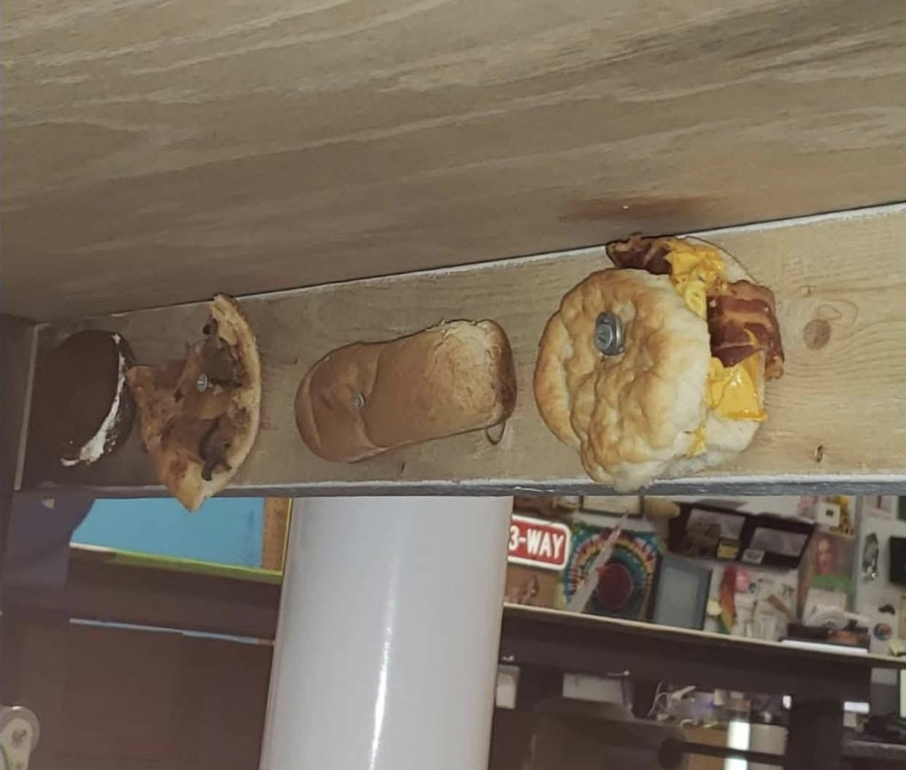 Three half-eaten burgers and sandwiches are stuck underneath a wooden table, with various objects and signs visible in the blurred background.