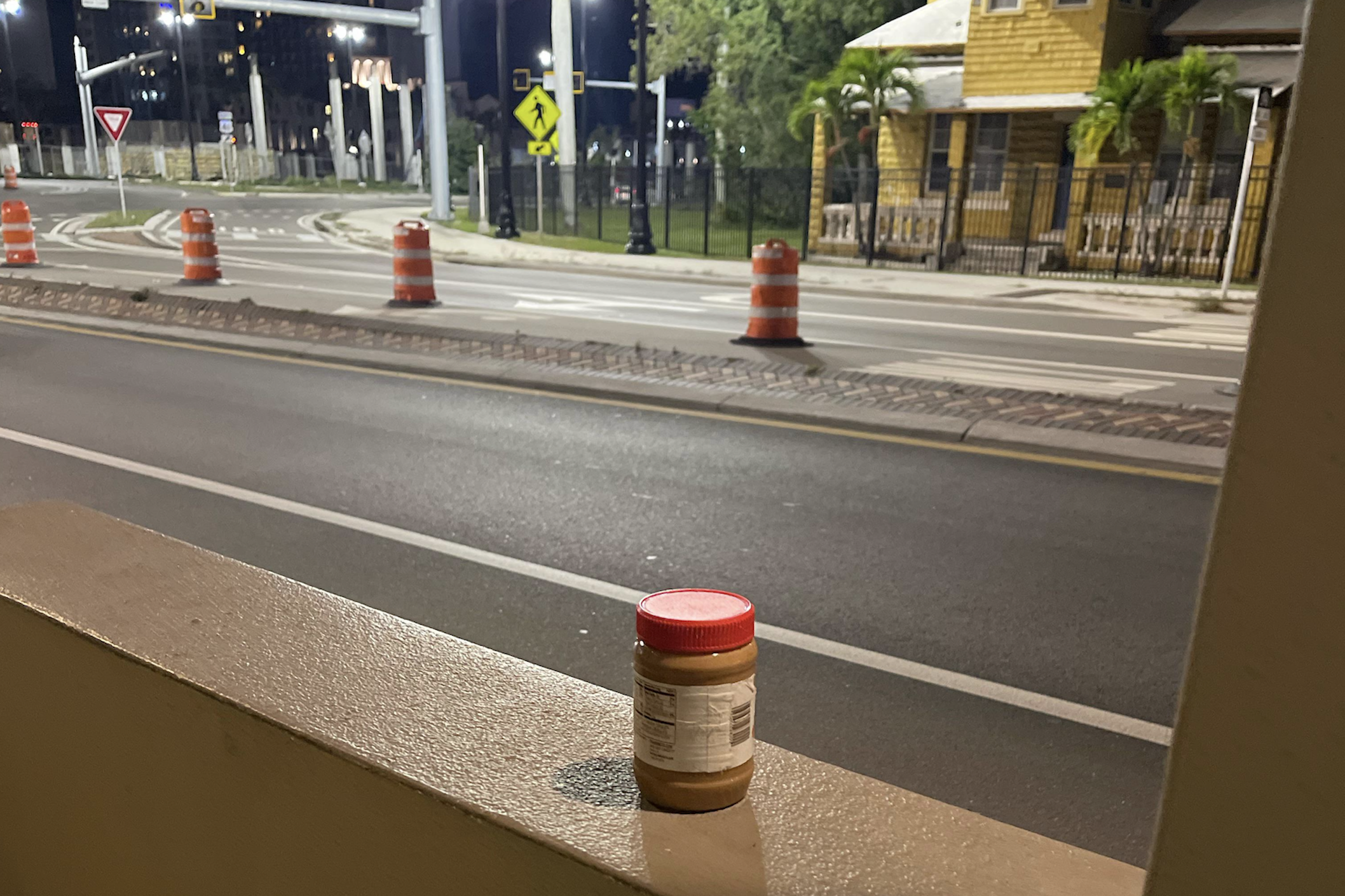 A small jar with a red lid sits on a low wall at night, overlooking an empty street with orange traffic barrels and a yellow house in the background.