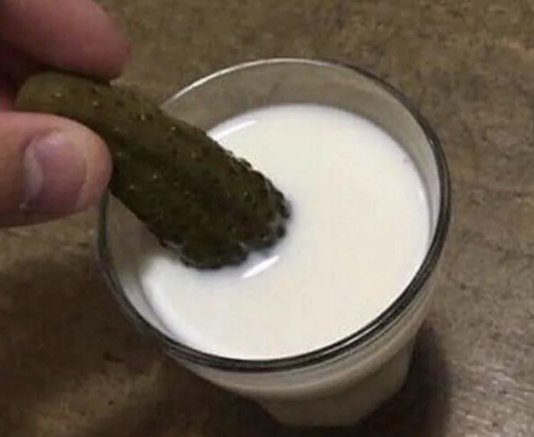 A hand dips a pickle into a clear glass filled with milk, set on a brown surface.