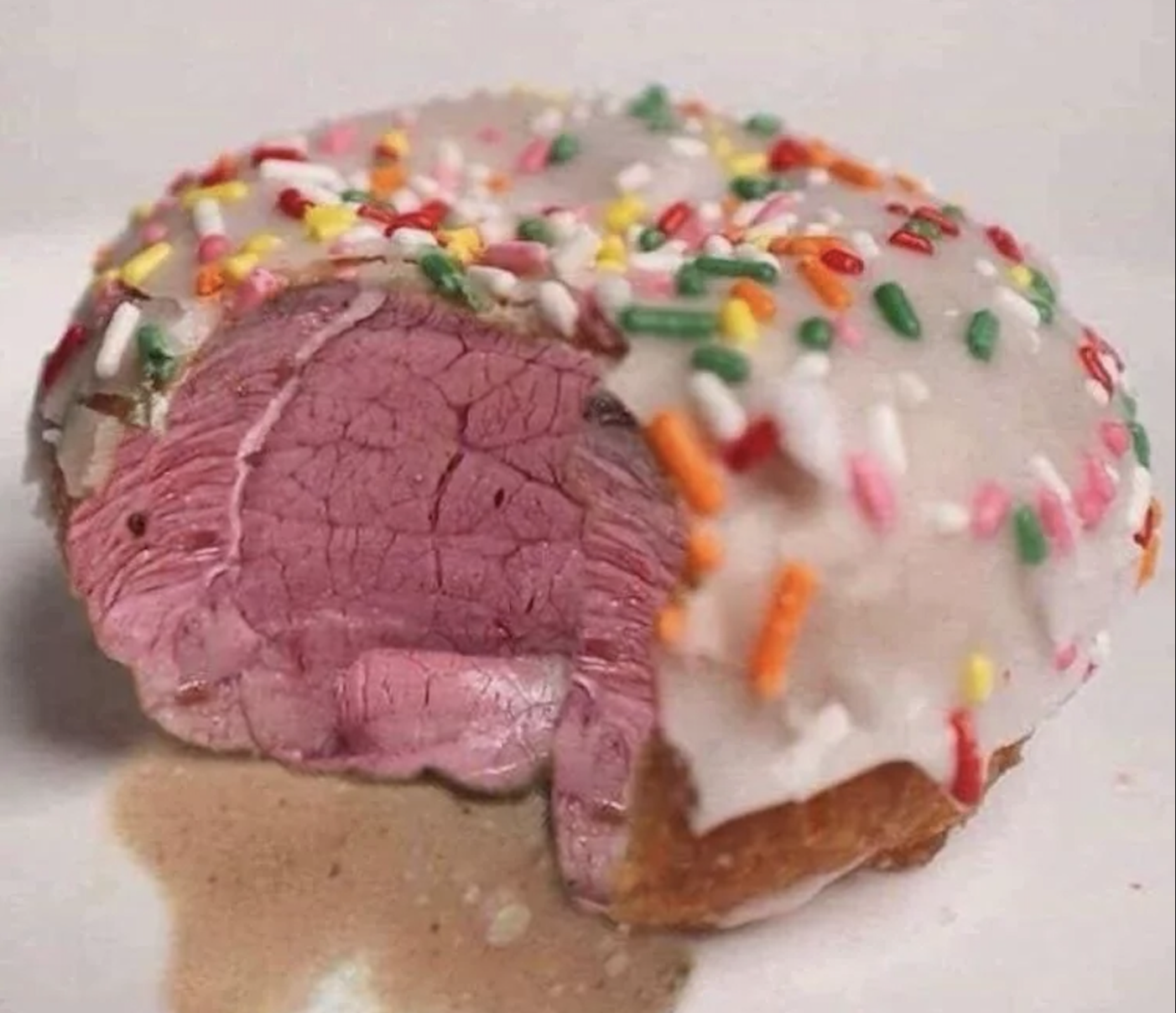A donut with white icing and colorful sprinkles, but the inside appears to be rare-cooked beef instead of regular donut bread. A bite has been taken out.
