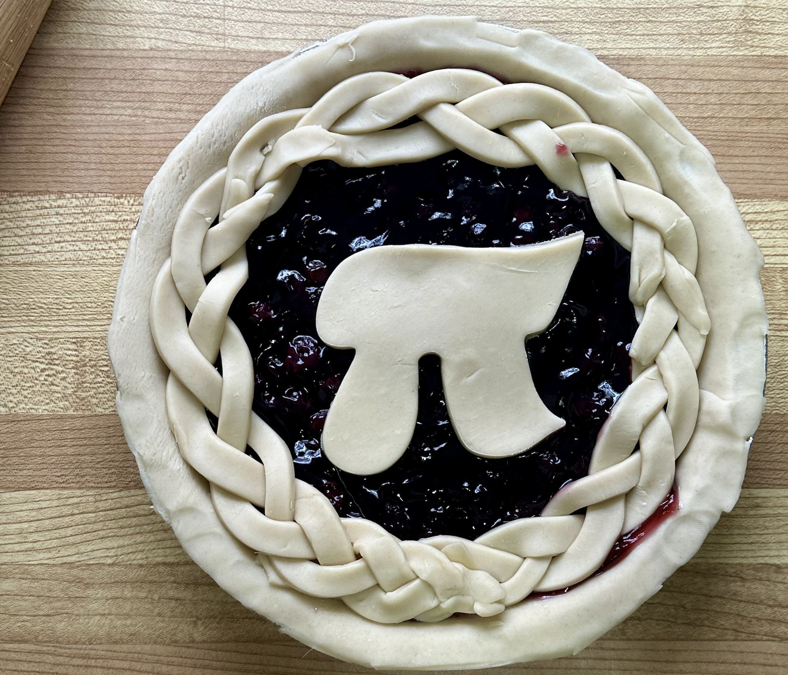 Unbaked berry pie topped with a braided crust edge and a large crust cutout shaped like the mathematical pi symbol in the center. The pie sits on a wooden surface.