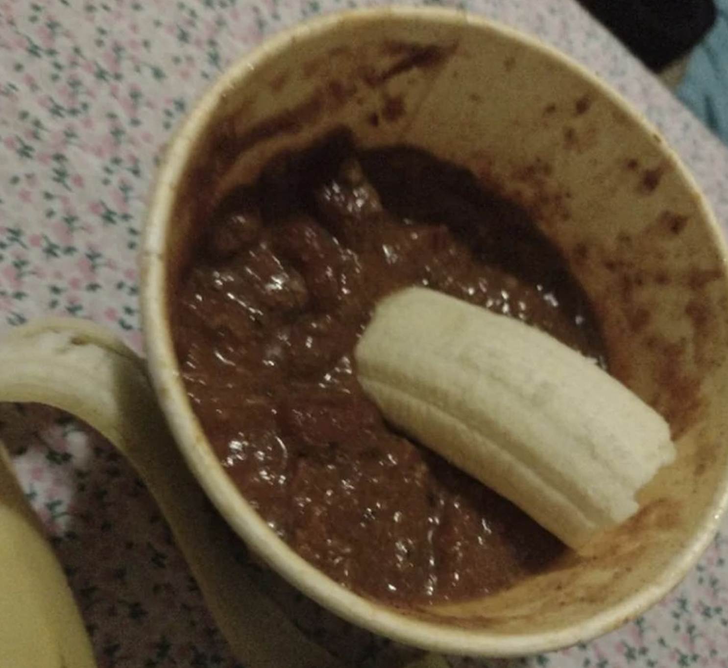 A mug filled with thick, dark stew or chili, with half of a peeled banana placed inside. The mug is on a surface covered by a pink and white floral-patterned fabric.