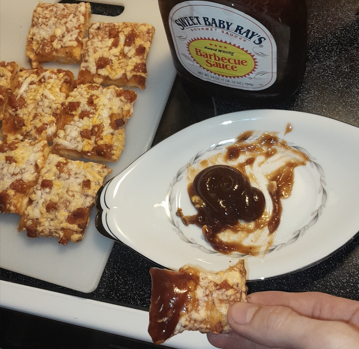 Hand holding a pizza snack dipped in barbecue sauce on a plate; more pizza snacks and a bottle of Sweet Baby Ray’s Barbecue Sauce are nearby on a white cutting board and stove.