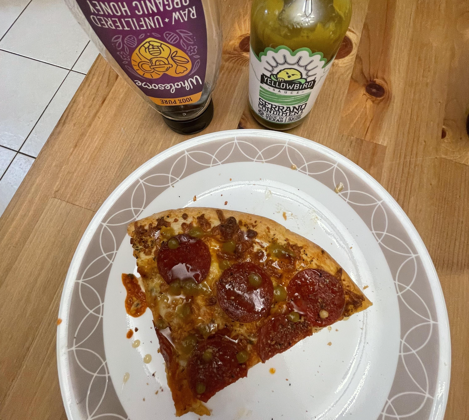 A plate with a slice of pepperoni pizza sits on a wooden table. Nearby are bottles of organic raw honey and Yellowbird Serrano hot sauce.