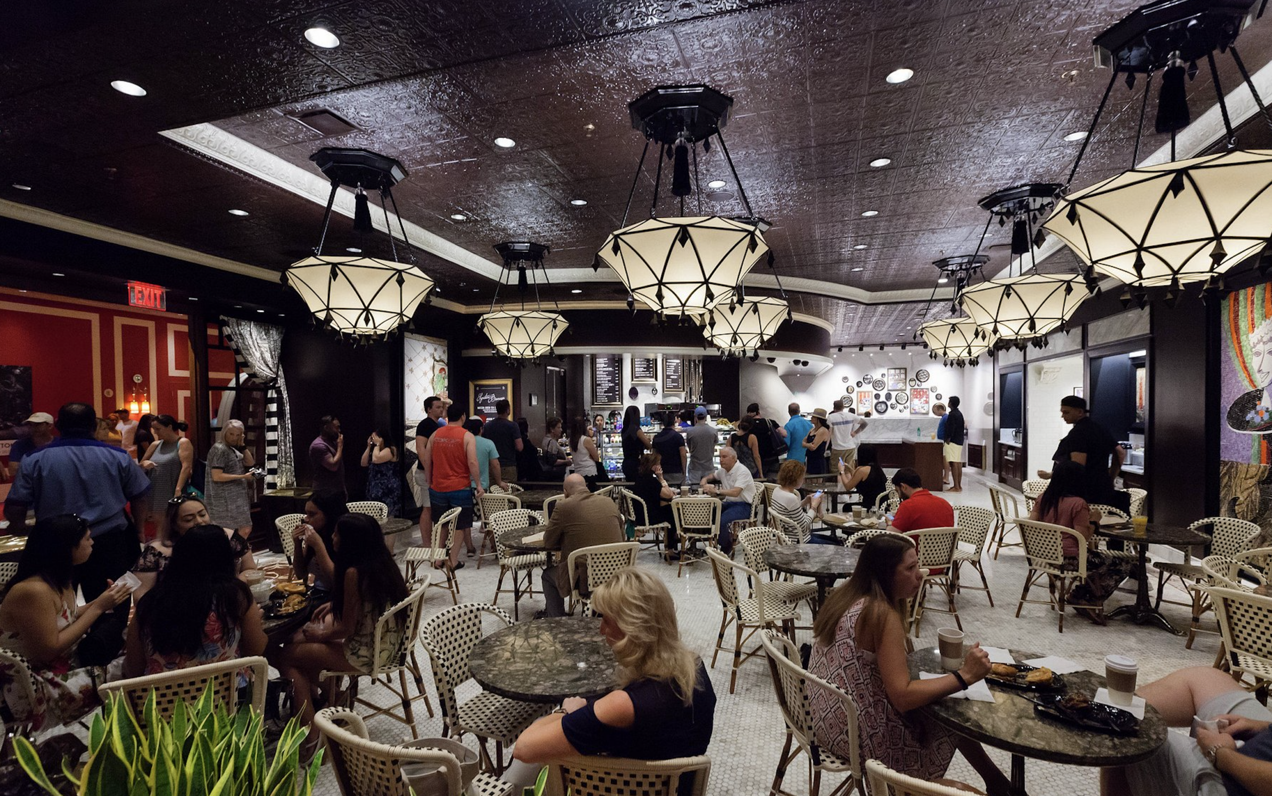 A busy cafe with round tables, patrons seated and eating, and people standing in line at the counter. The room has decorative light fixtures, a dark ceiling, and colorful wall art in the background.