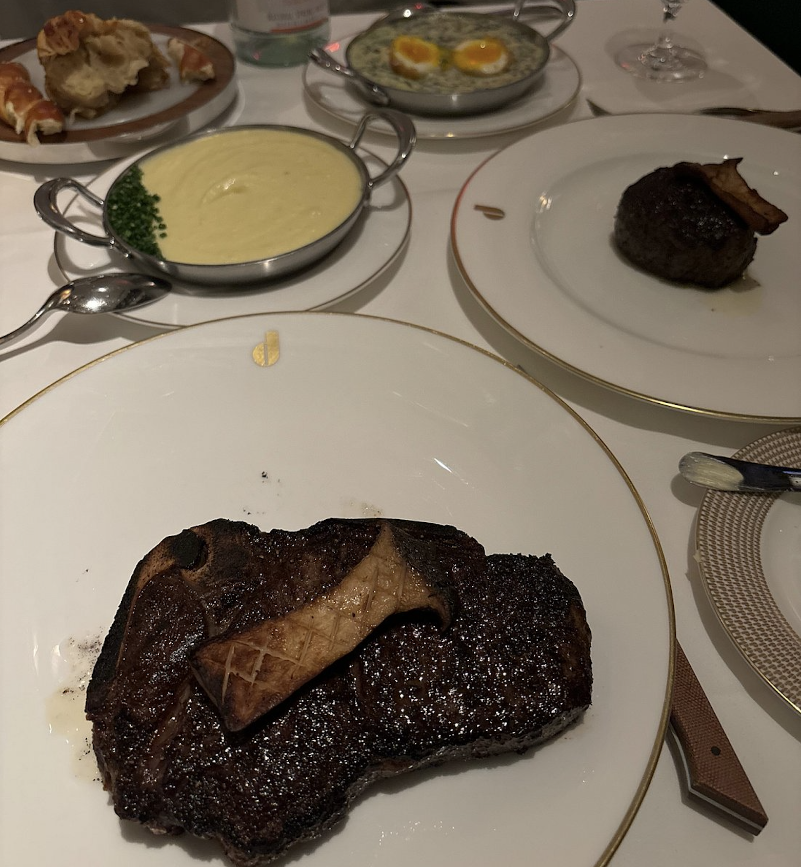 A table set with grilled steak topped with mushroom, mashed potatoes, creamed spinach with eggs, bread rolls, and another steak. White tablecloth and elegant dinnerware suggest a fine dining setting.