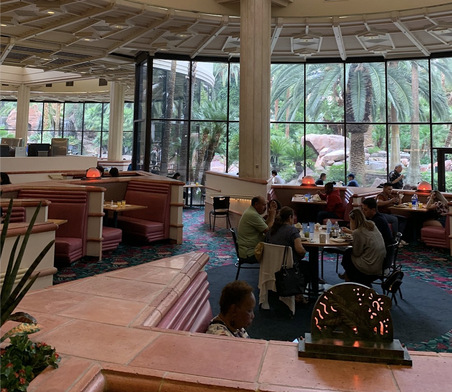 Diners sit at tables and pink booths inside a restaurant with large windows overlooking palm trees. Some people eat, talk, or look at menus. The space is brightly lit with red lamps and lush greenery outside.