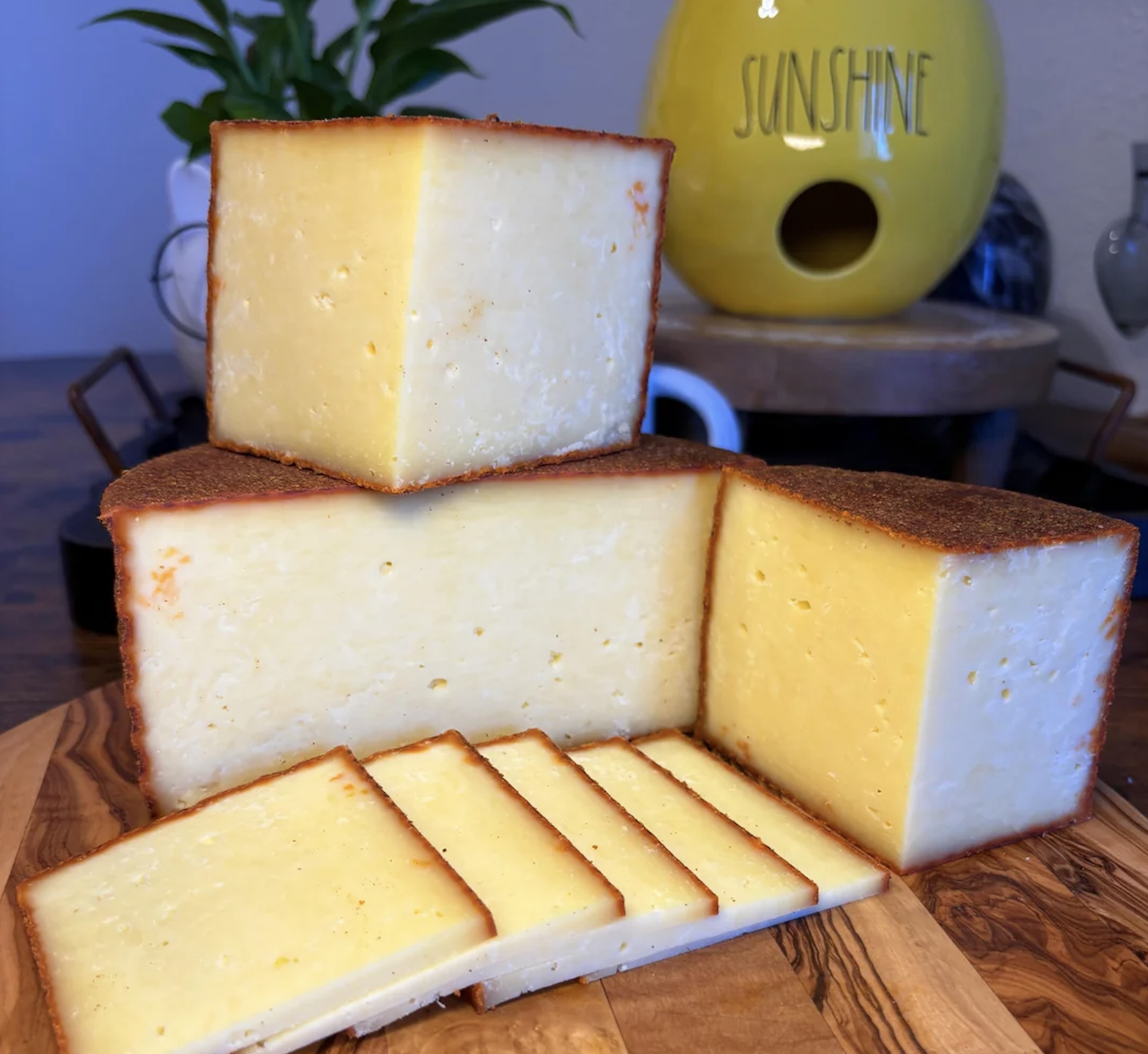 Several thick blocks and thin slices of yellow cheese with a brown rind are arranged on a wooden board. In the background, there’s a yellow container labeled “SUNSHINE” and a green plant.
