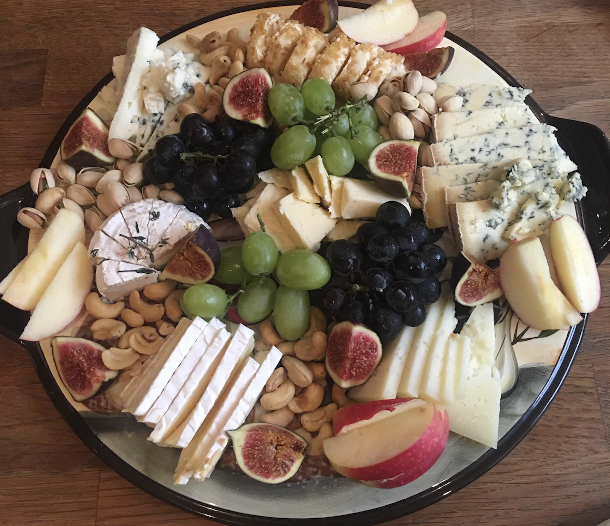 A platter with assorted cheeses, figs, grapes, apple slices, pistachios, and cashews, arranged on a round plate set on a wooden table.