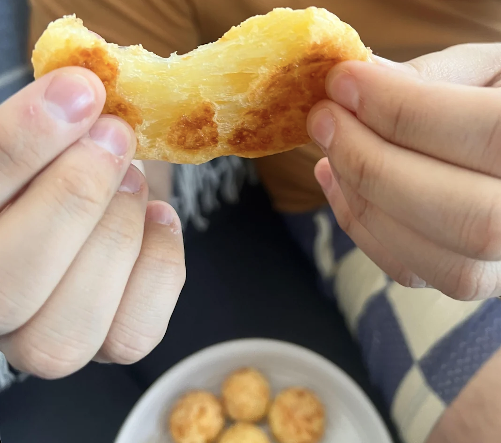 Two hands hold and stretch a cheesy, golden-brown baked snack above a white bowl containing several more snacks. The background includes a person’s lap and part of a blanket.