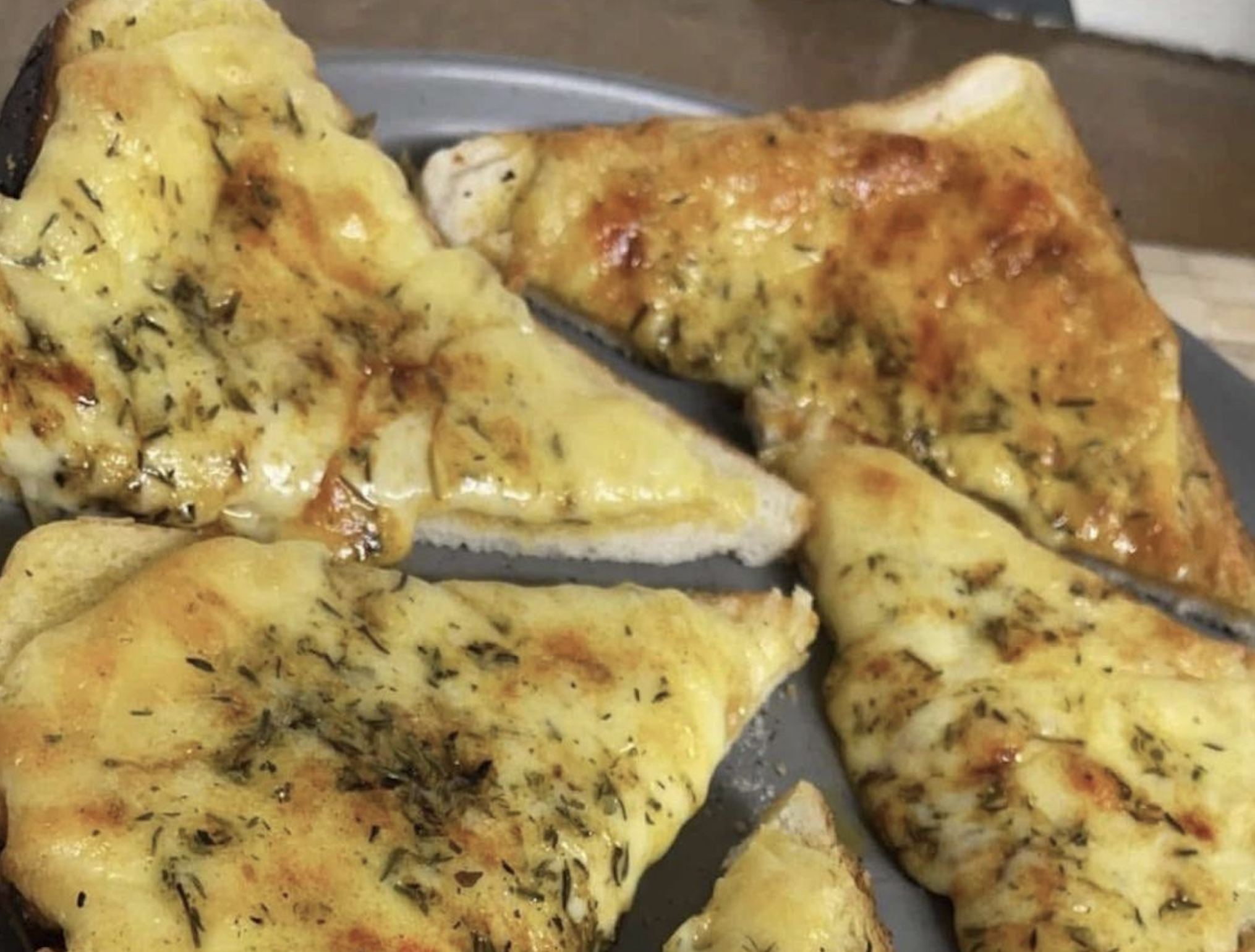 Four slices of cheesy garlic bread with herbs on a grey plate. The bread looks toasted, with melted cheese and sprinkled seasoning visible on top.