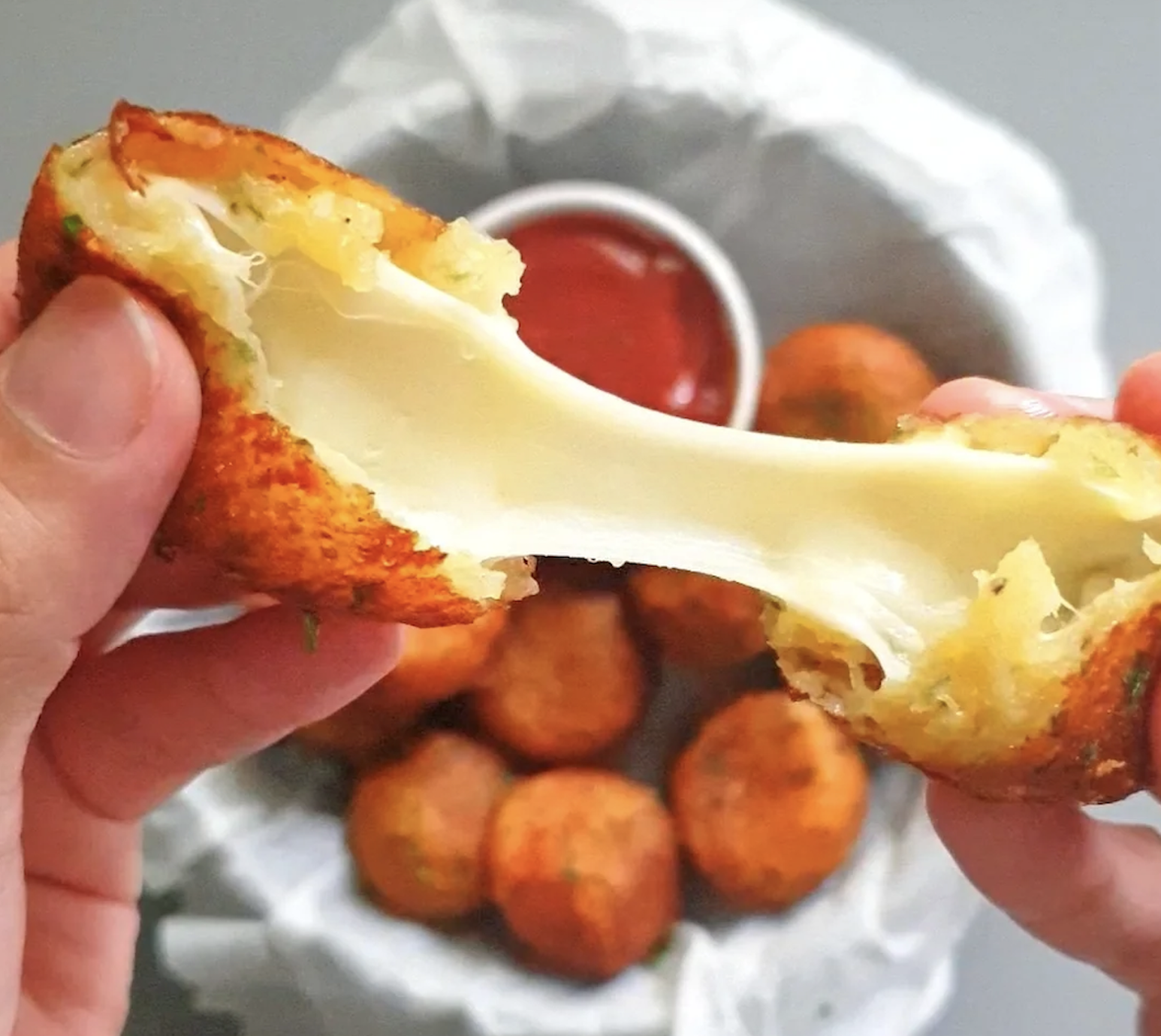 Hands pull apart a golden, crispy cheese ball with gooey melted cheese stretching between the halves. In the background, more cheese balls and a small dish of red sauce sit in a white-lined bowl.