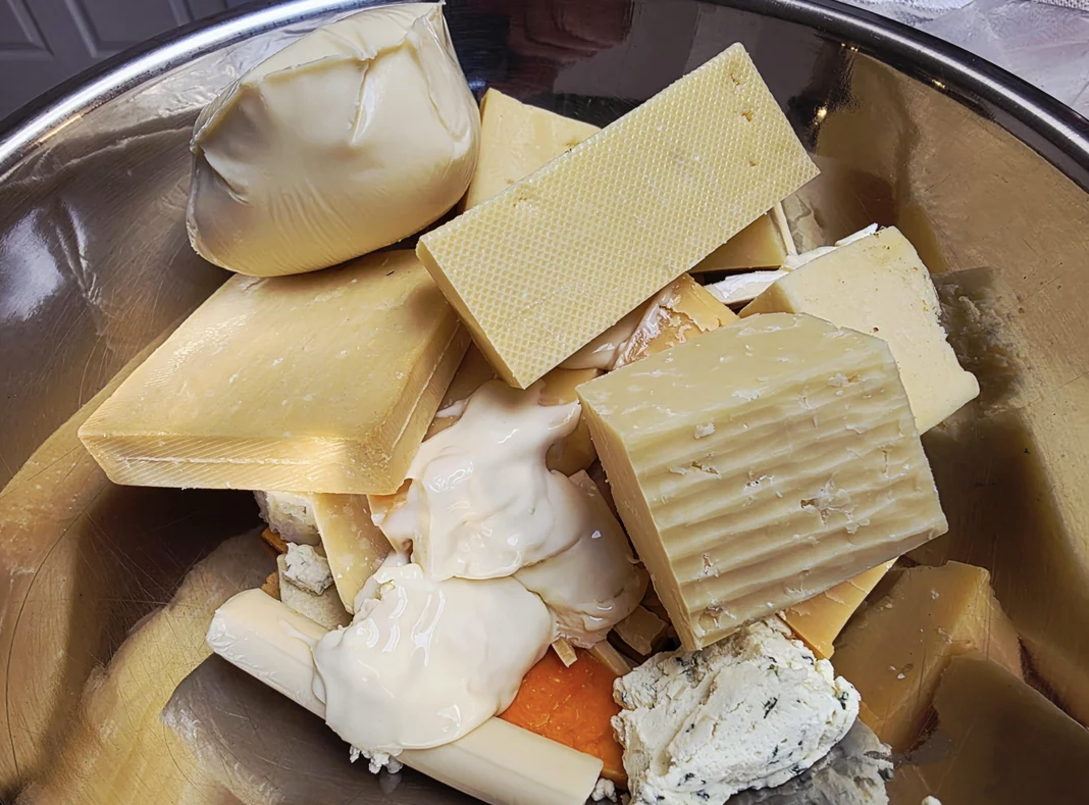 A stainless steel bowl filled with assorted cheese blocks, creamy cheeses, and soft cheese pieces, all piled together in various shapes and textures.