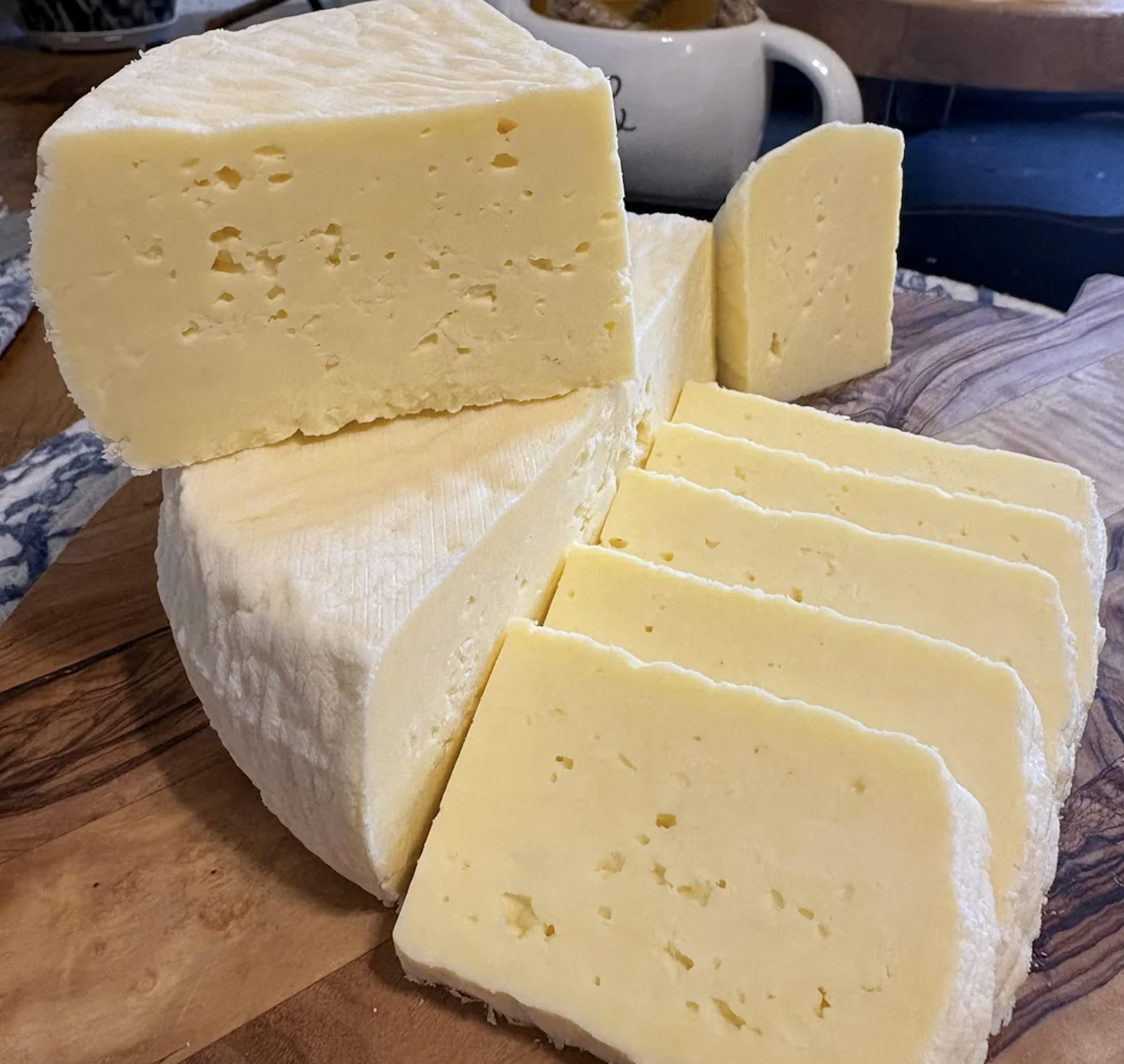 A block of semi-soft, pale yellow cheese is partially sliced, with several even slices arranged on a wooden board. The cheese has a smooth, slightly crumbly texture and a white rind.