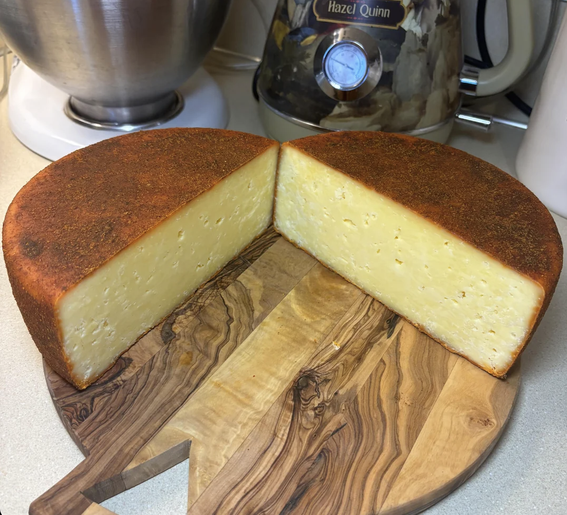 A round wheel of cheese with a reddish rind is cut in half on a wooden board. The creamy yellow interior is firm and smooth. Kitchen appliances are visible in the background.