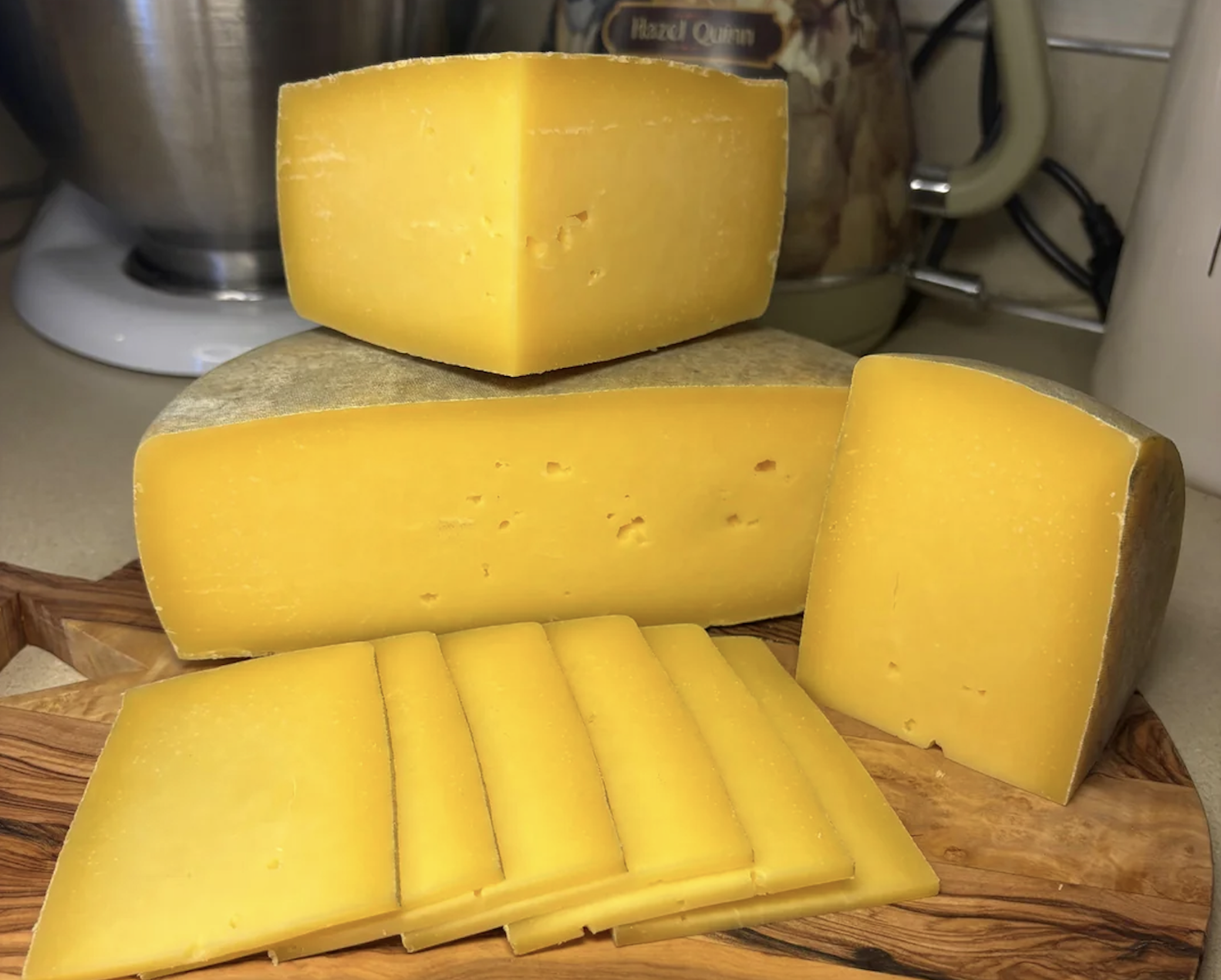 A block of yellow cheese is sliced on a wooden cutting board, with wedges and several thin rectangular slices arranged in front, set on a kitchen counter with appliances in the background.