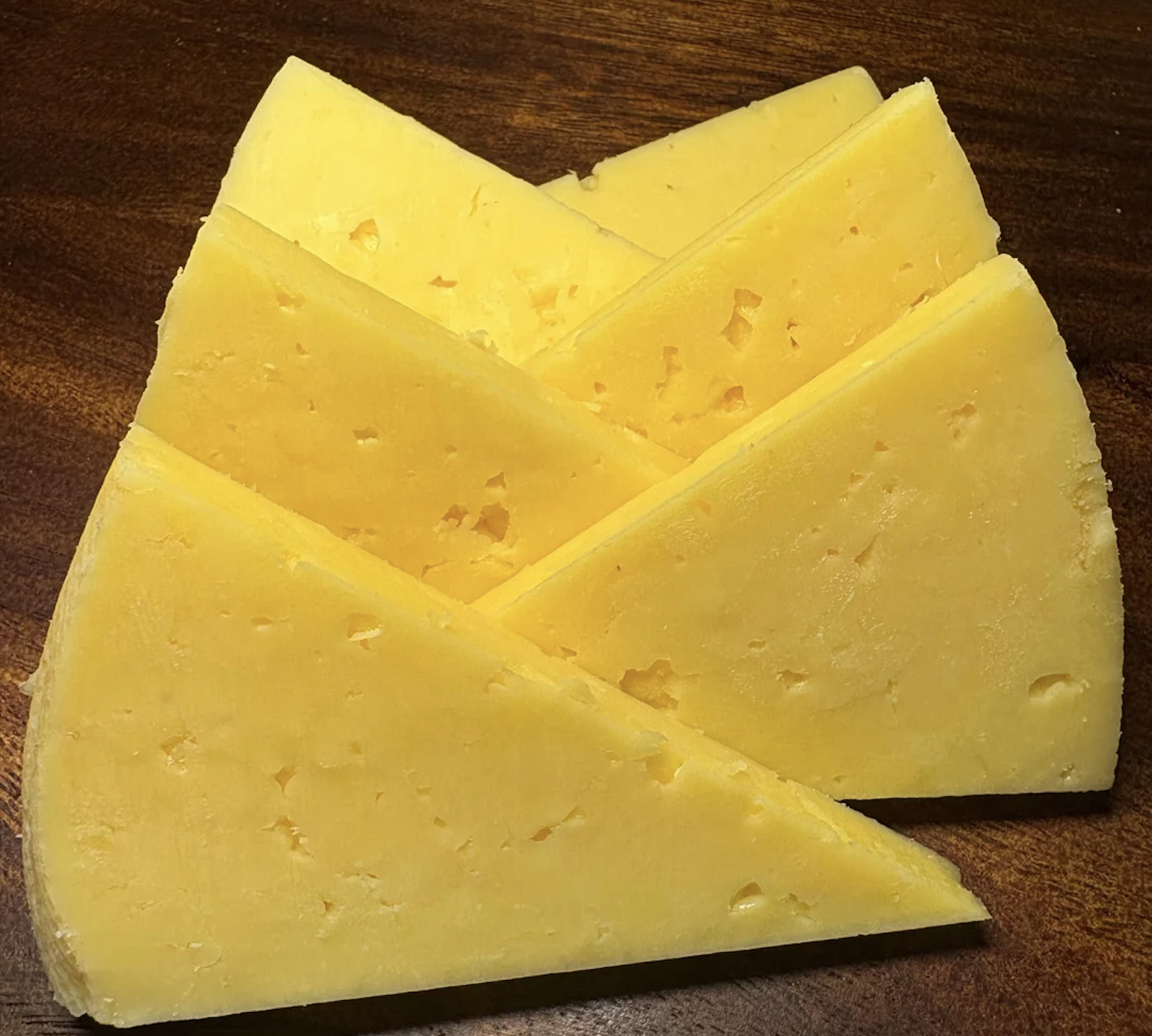 Five triangular slices of yellow cheese are arranged on a wooden surface, overlapping in a fan-like pattern. The cheese has a smooth texture with small holes and a firm appearance.