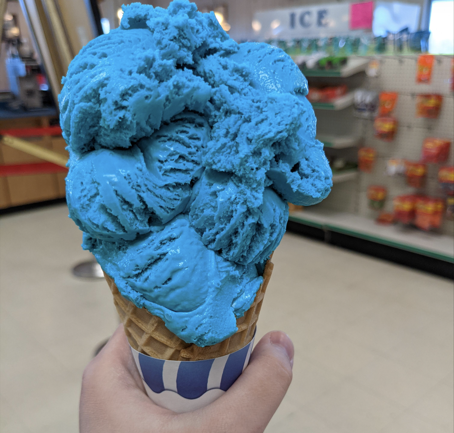 A hand holds a waffle cone filled with bright blue ice cream inside a store, with shelves and a sign reading “ICE” visible in the background.