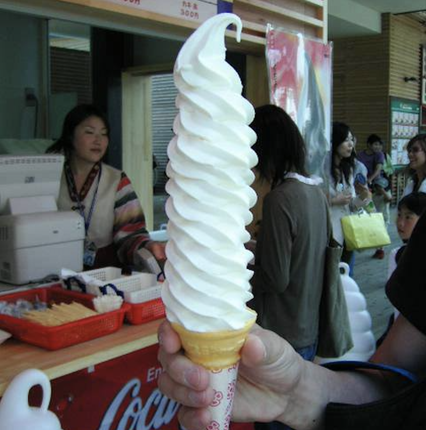 A person holds a very tall soft-serve ice cream cone in front of a counter, with a vendor and several people in the background at an indoor market or shop.