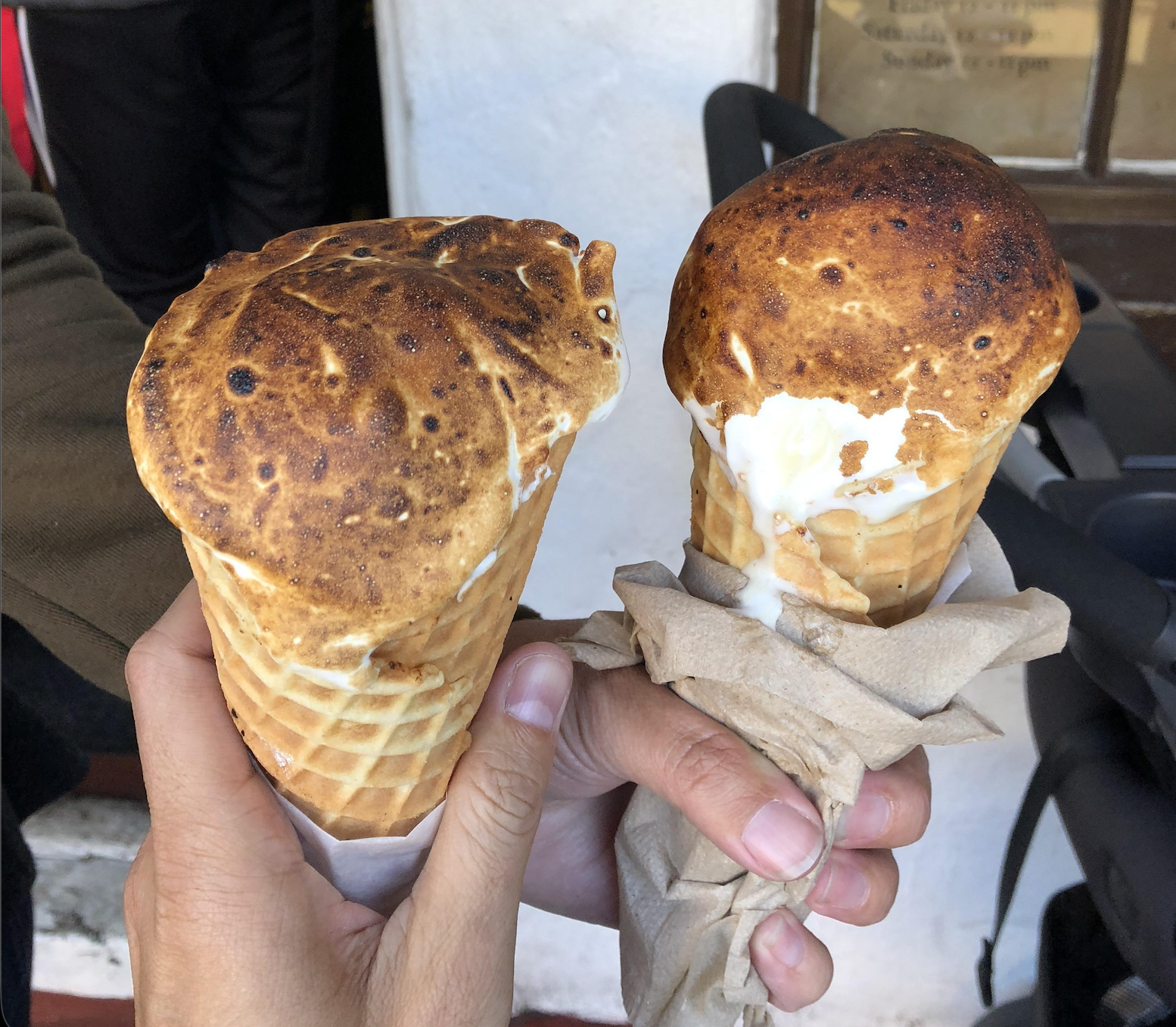 Two hands holding waffle cones with ice cream topped by a layer of toasted, golden brown marshmallow, partially wrapped in napkins.