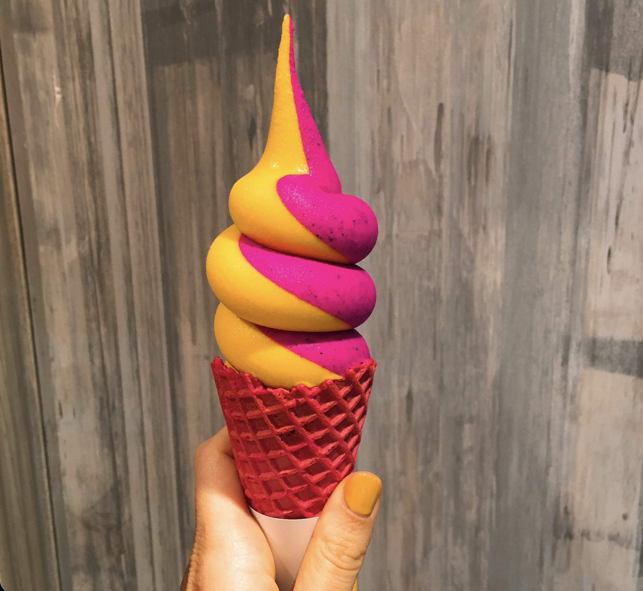 A hand holds a vibrant ice cream cone with yellow and magenta swirled soft serve in a pink waffle cone, matching the nail polish. The background is a textured gray wall.
