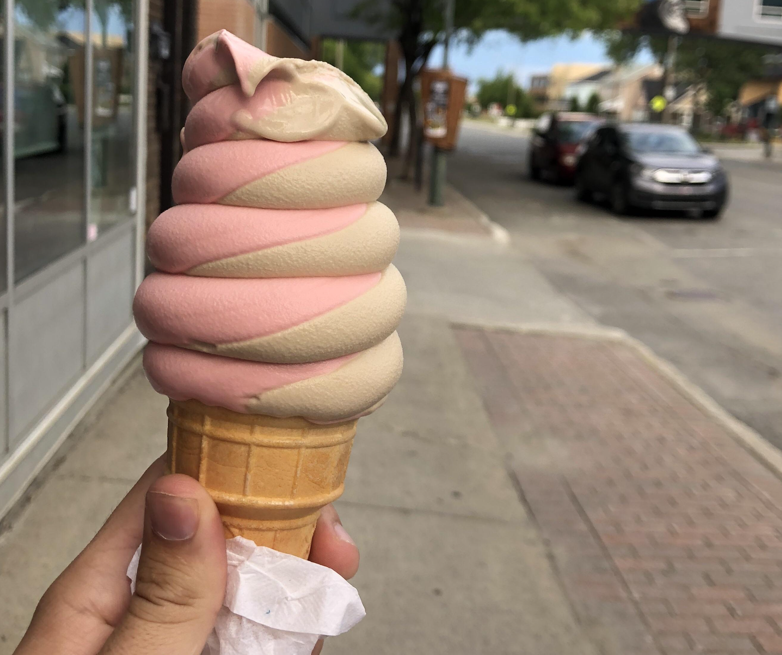 A hand holds a swirled pink and beige ice cream cone on a city sidewalk. The cone is wrapped with a napkin, and a street with cars and trees is visible in the background.