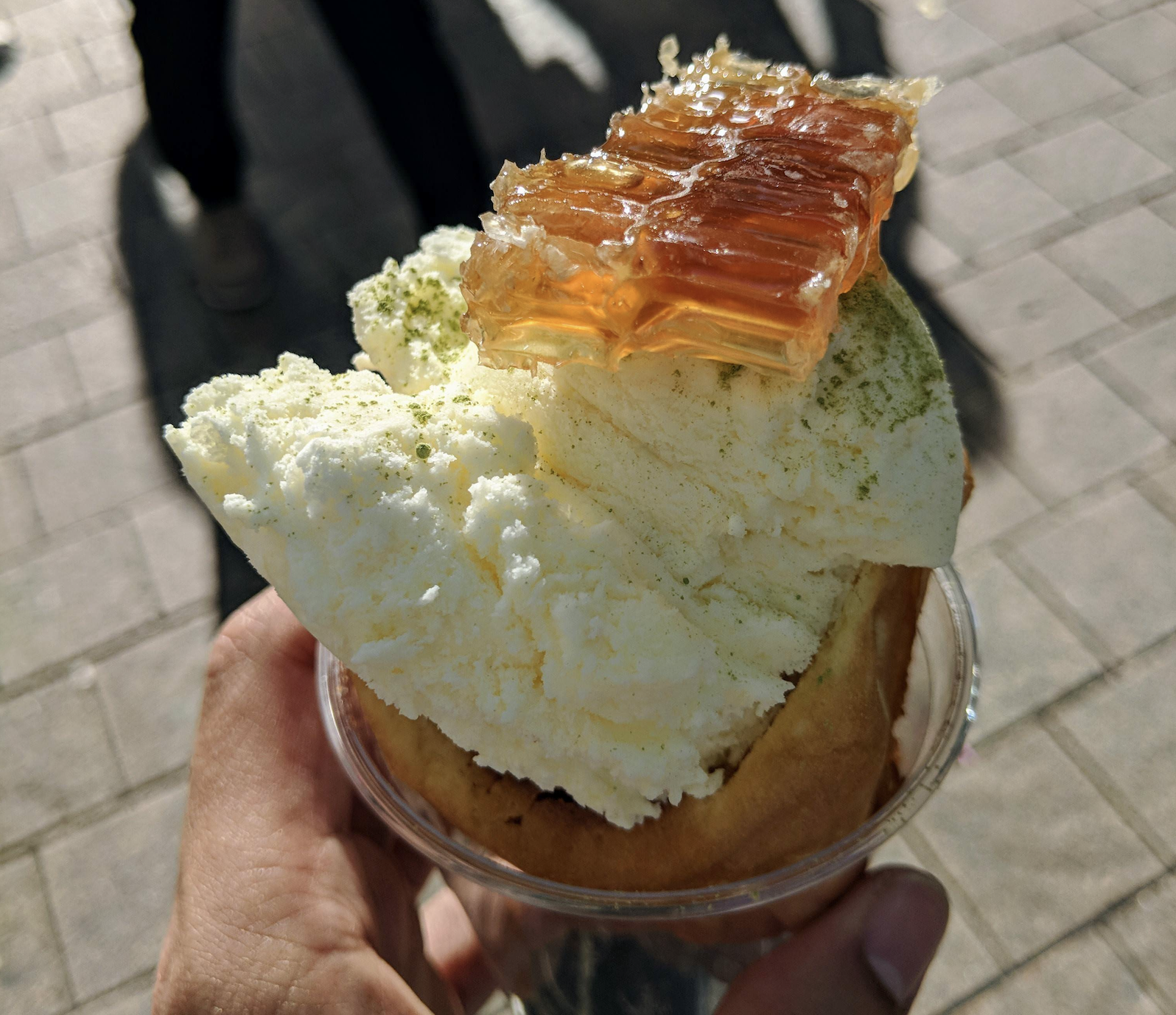 A hand holds a dessert with vanilla ice cream, a piece of honeycomb, and green powder, served in a pastry cup, with a sunlit street background.