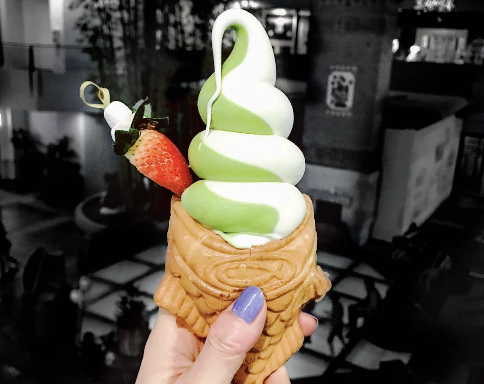 A hand with purple nail polish holds a fish-shaped waffle cone filled with green and white swirled soft serve ice cream, topped with a strawberry and white chocolate.