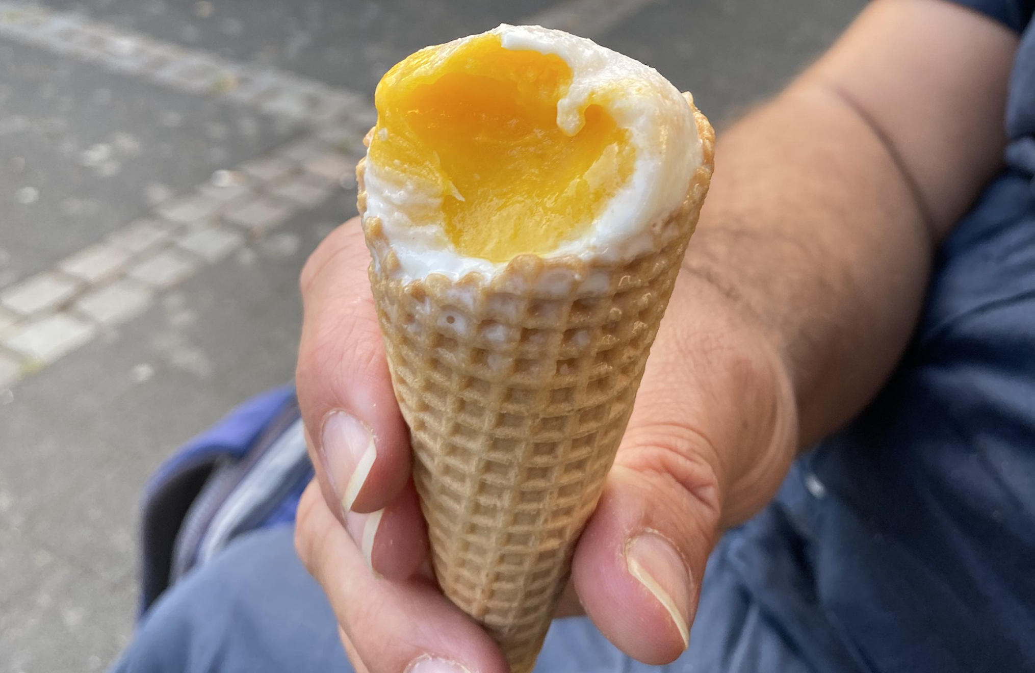 A hand holds an ice cream cone with a bite taken out, revealing yellow and white ice cream inside. The background is slightly blurred pavement.