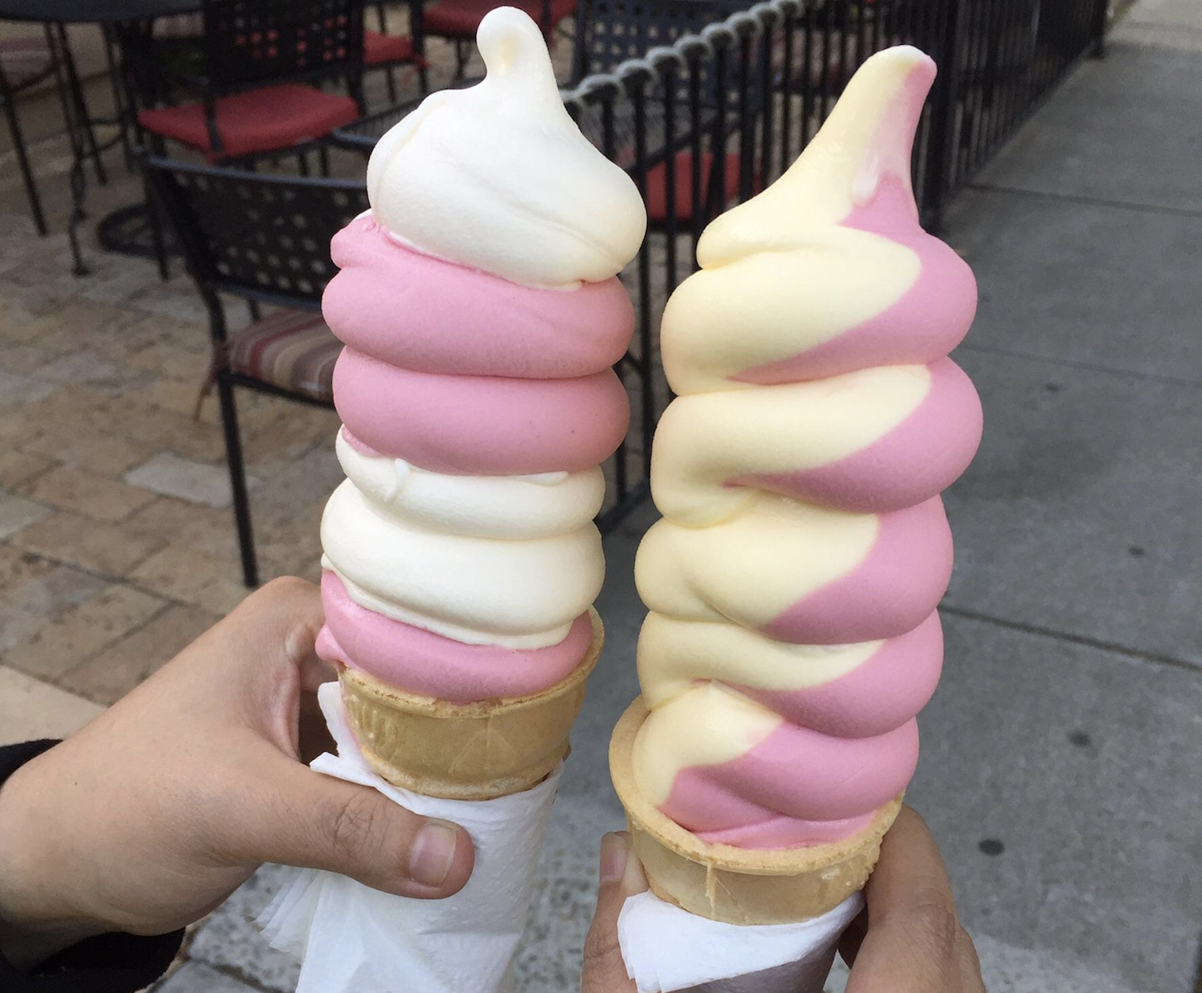 Two hands holding soft serve ice cream cones, each with swirled pink and white or pink and yellow ice cream, over a sidewalk and outdoor seating area.