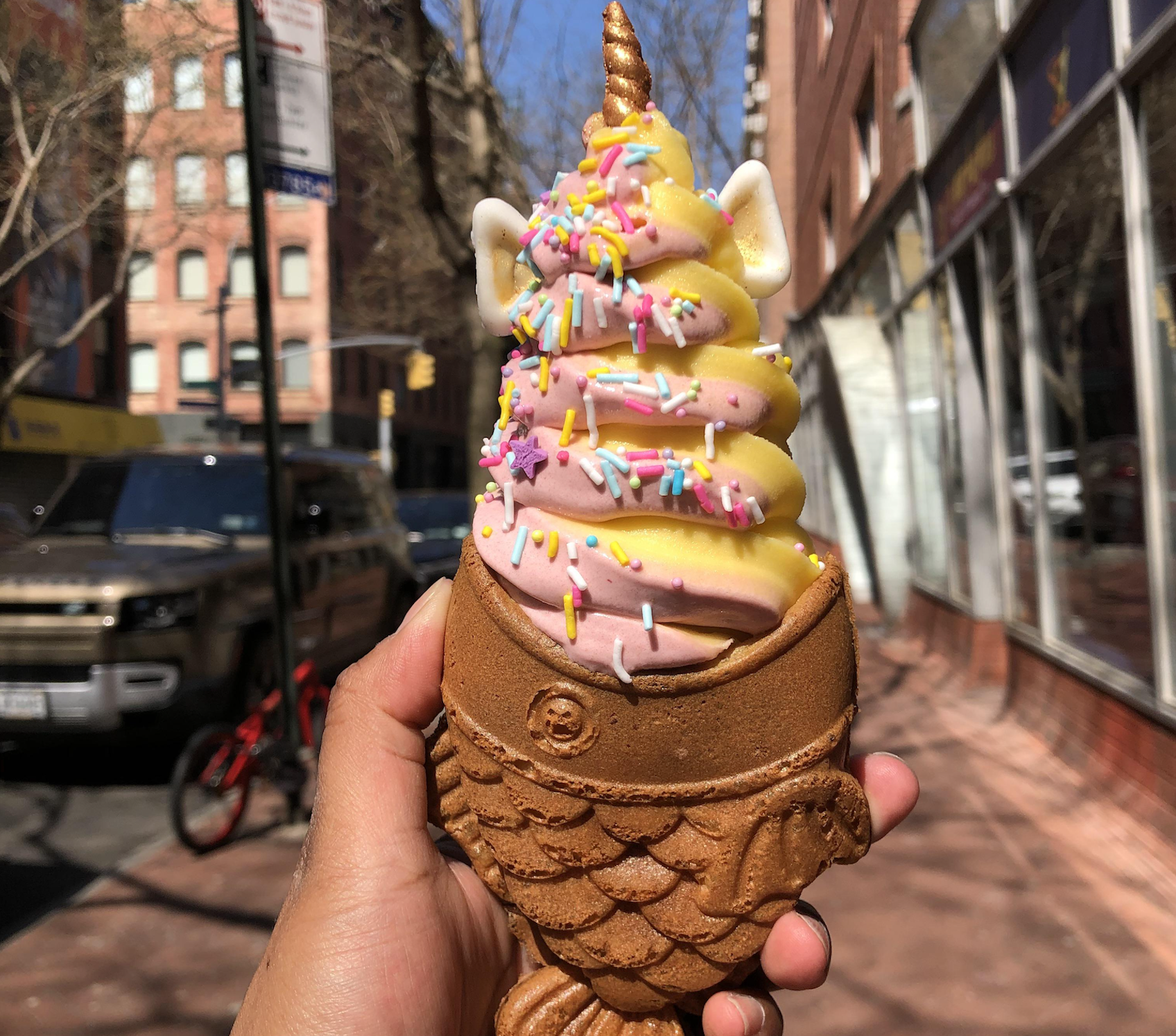 A hand holds a taiyaki cone filled with swirled pink and yellow unicorn-themed soft serve topped with rainbow sprinkles, with a city street in the background.