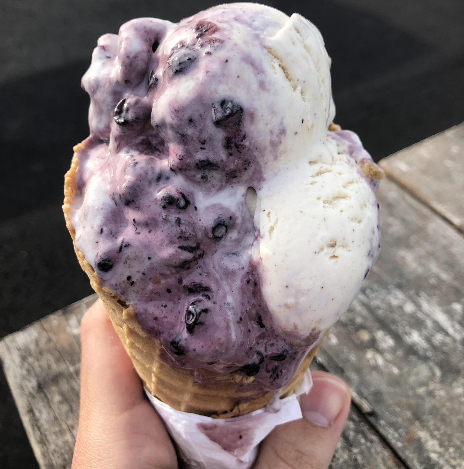 A hand holding an ice cream cone with two scoops, one purple with berries and the other white, over a wooden table with a dark background.